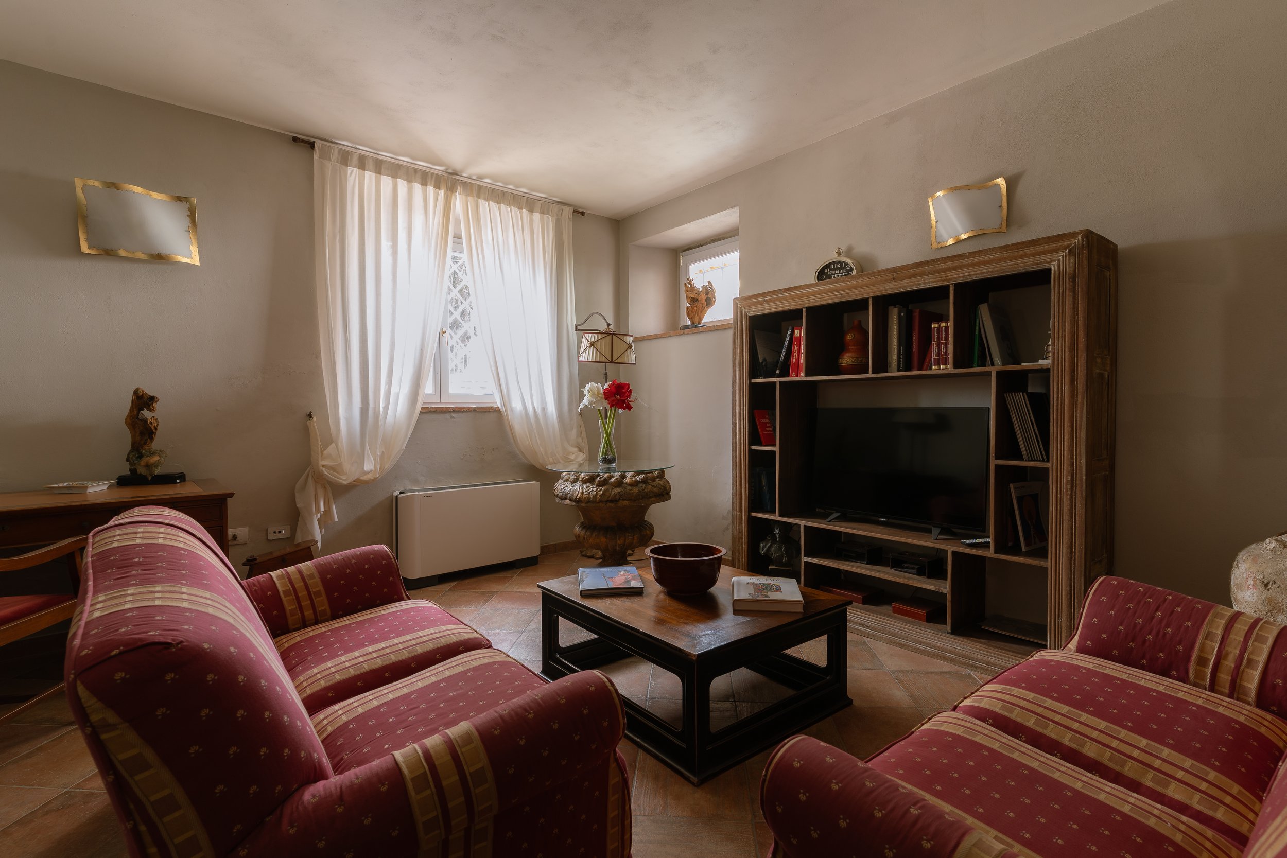 Living room with two patterned pink sofas, a wooden coffee table with books and a bowl, a wooden entertainment center with a TV, a window with white curtains, a small table with a vase and flowers, and various decorative items on shelves and walls.