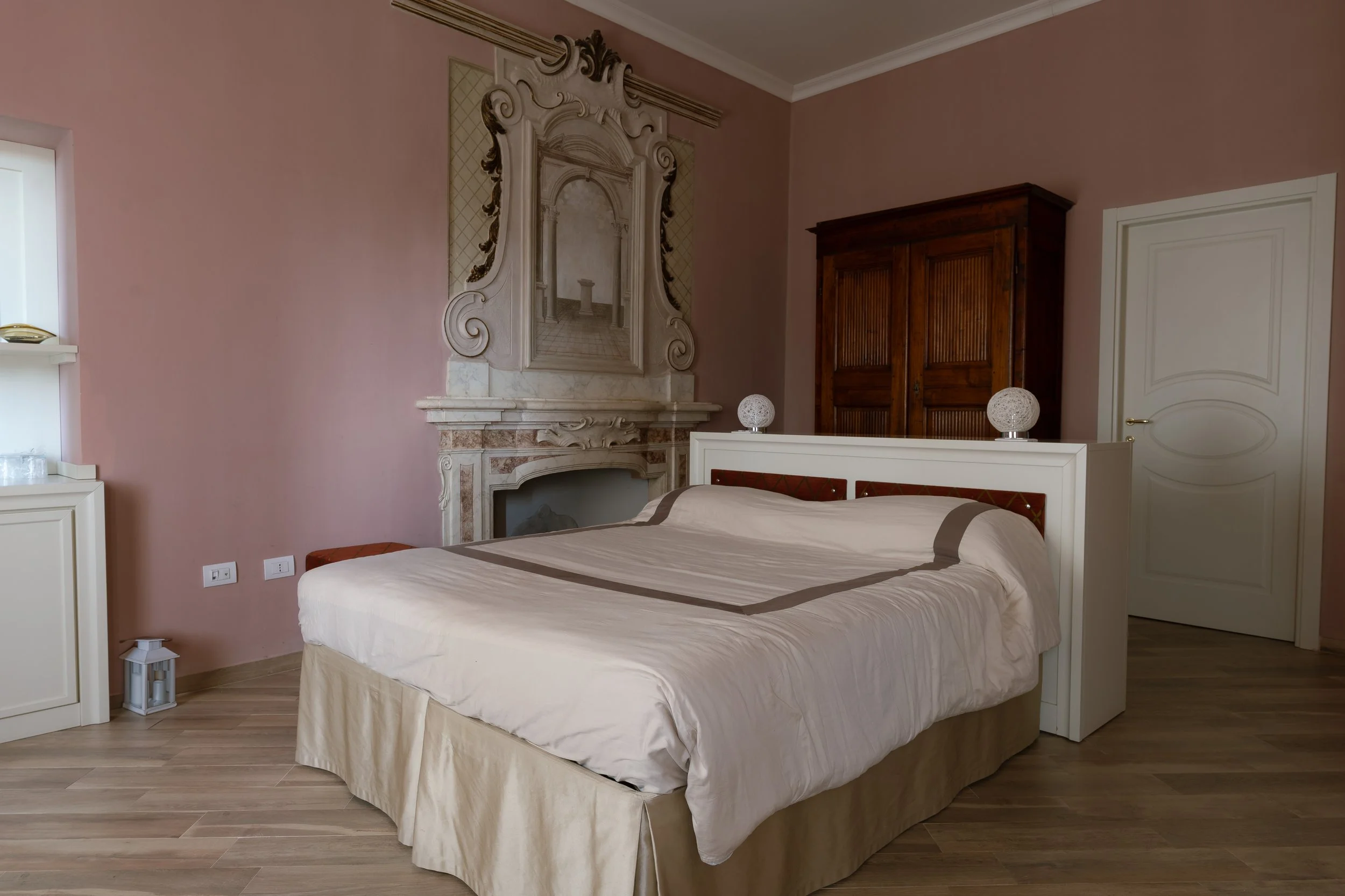 A bedroom with a beige bed, a wooden wardrobe, a white fireplace, and pink walls.