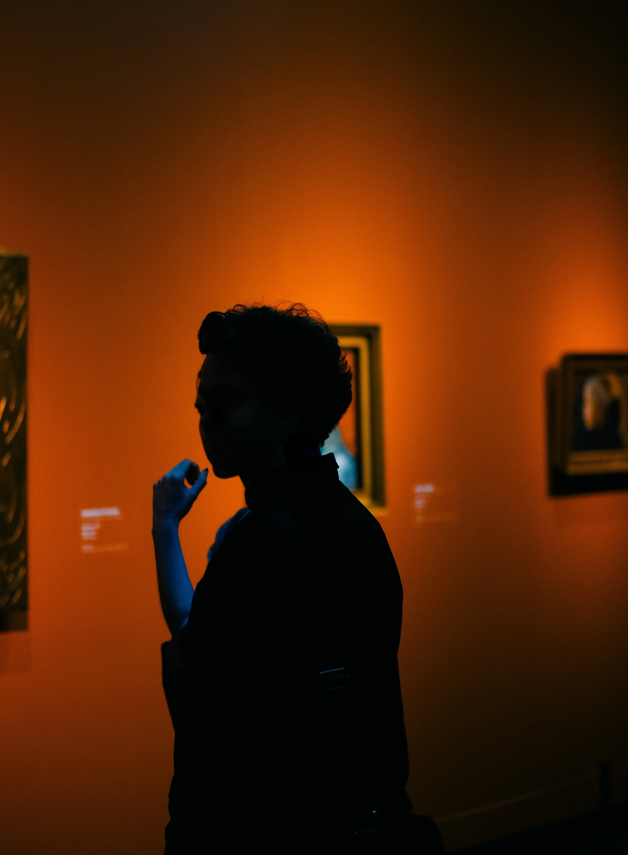 Silhouette of a person with styled hair, standing in front of an orange-lit gallery wall with framed artwork.