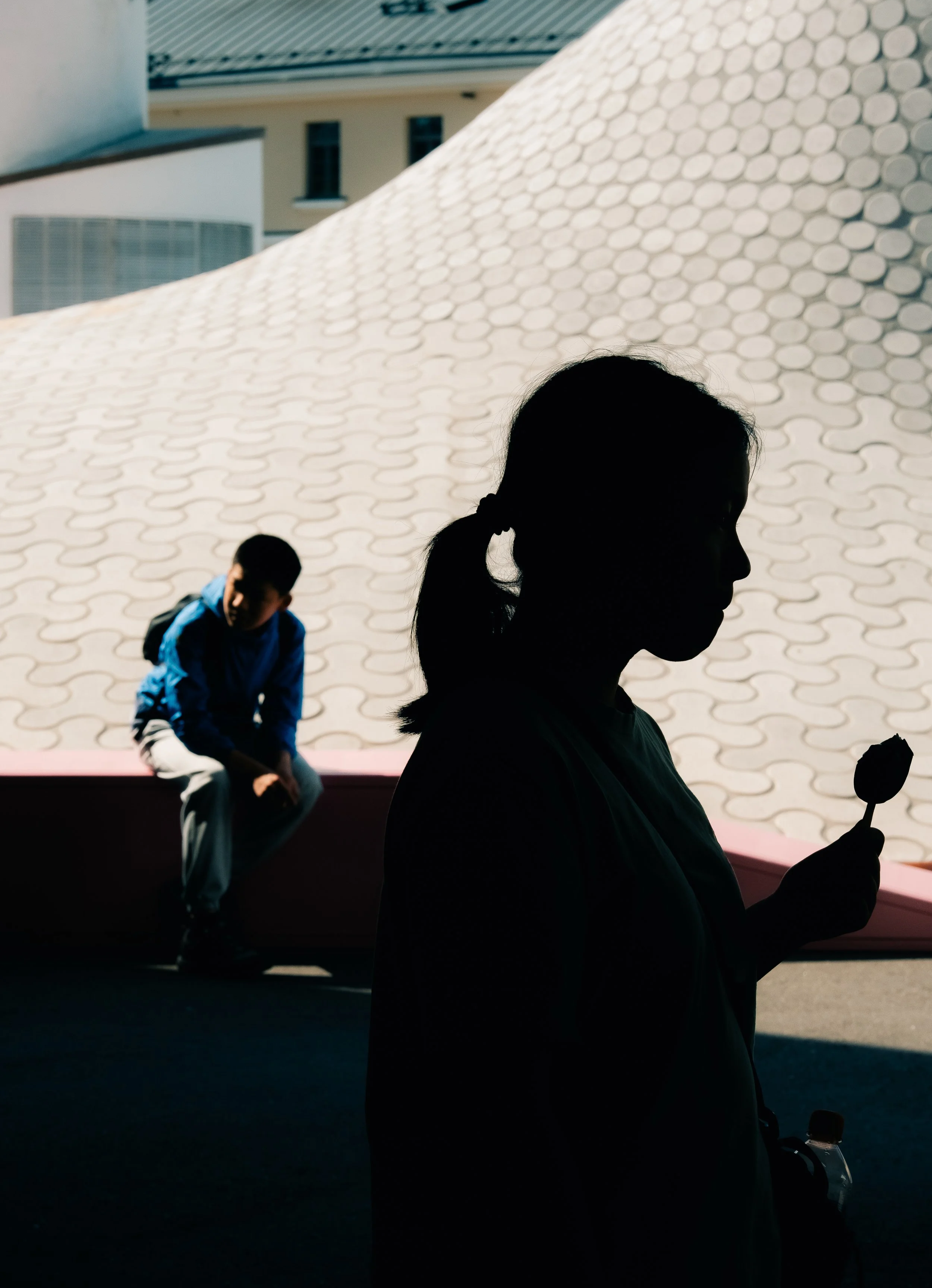 A silhouette of a woman holding a popsicle with a young boy sitting on a ledge in the background, with sunlight casting shadows on a patterned wall.