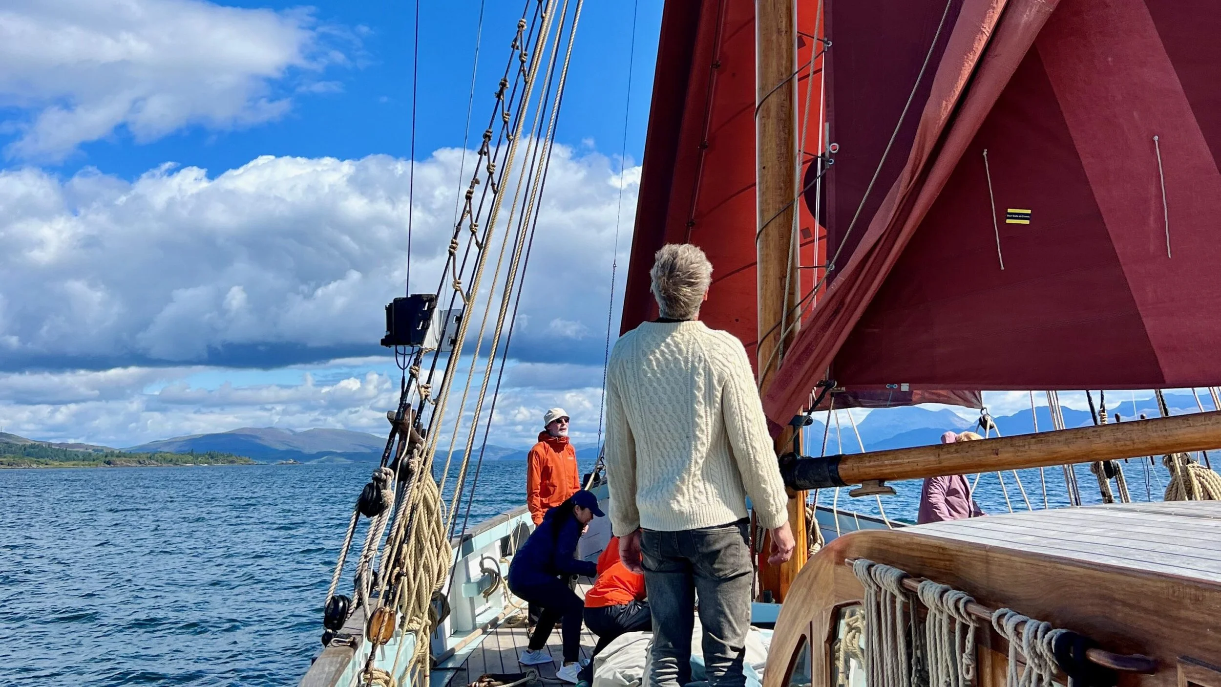 People on a sailboat with red sails on a body of water, mountains in the distance, and partly cloudy skies overhead.
