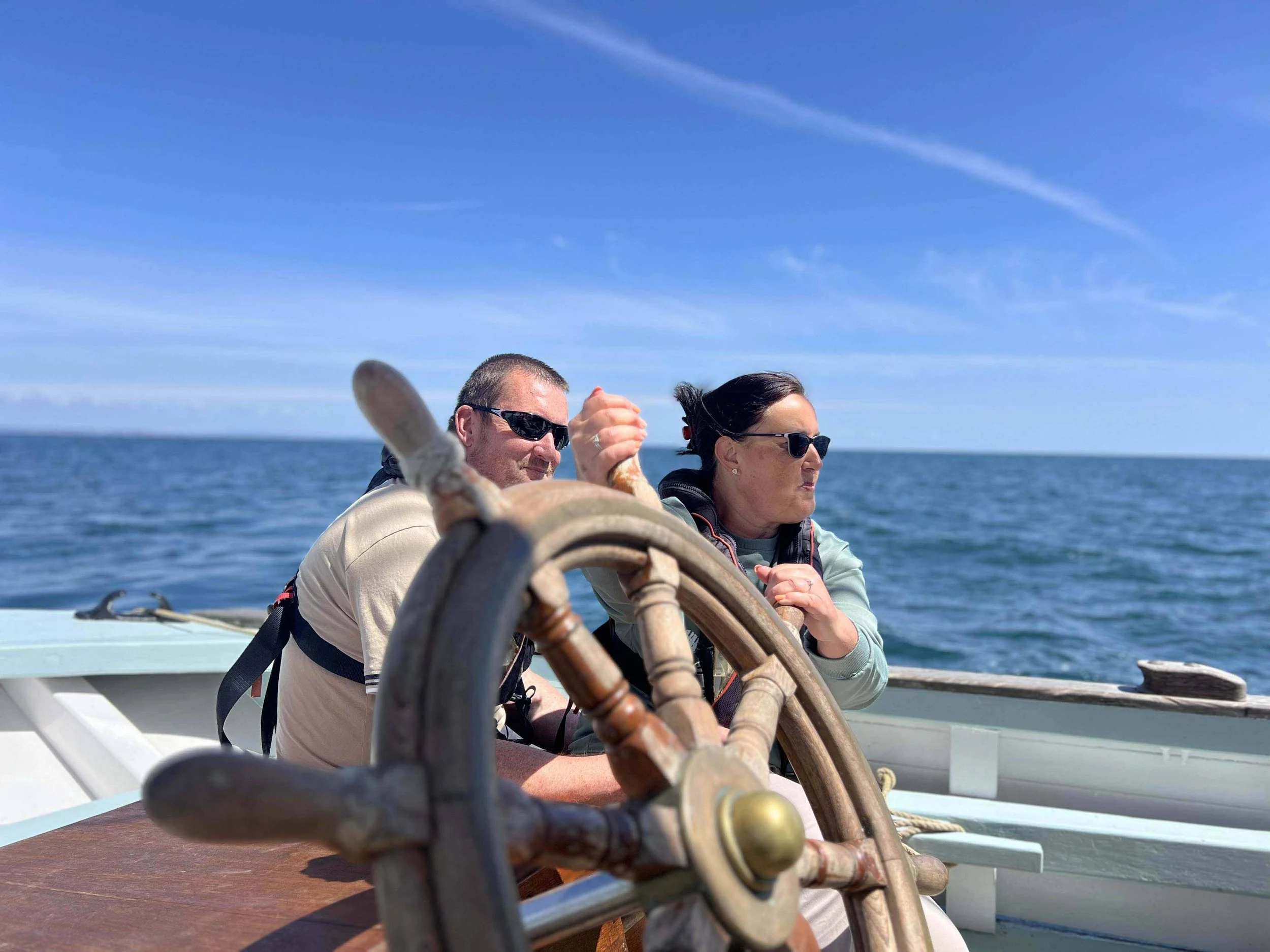 Two people on Provident, a man and a woman, both wearing sunglasses, are steering the boat. The man is holding the Provident wheel, and the woman looks forward with a serious expression. The ocean and a clear blue sky are in the background.