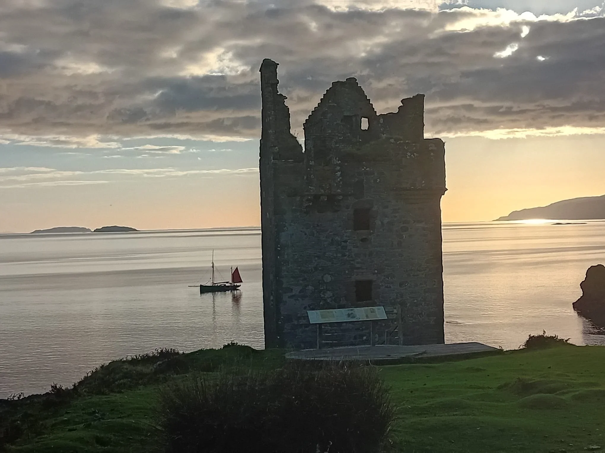 A silhouette of a ruined stone tower by the water at sunset, with cloudy sky and a boat with red sails in the background.
