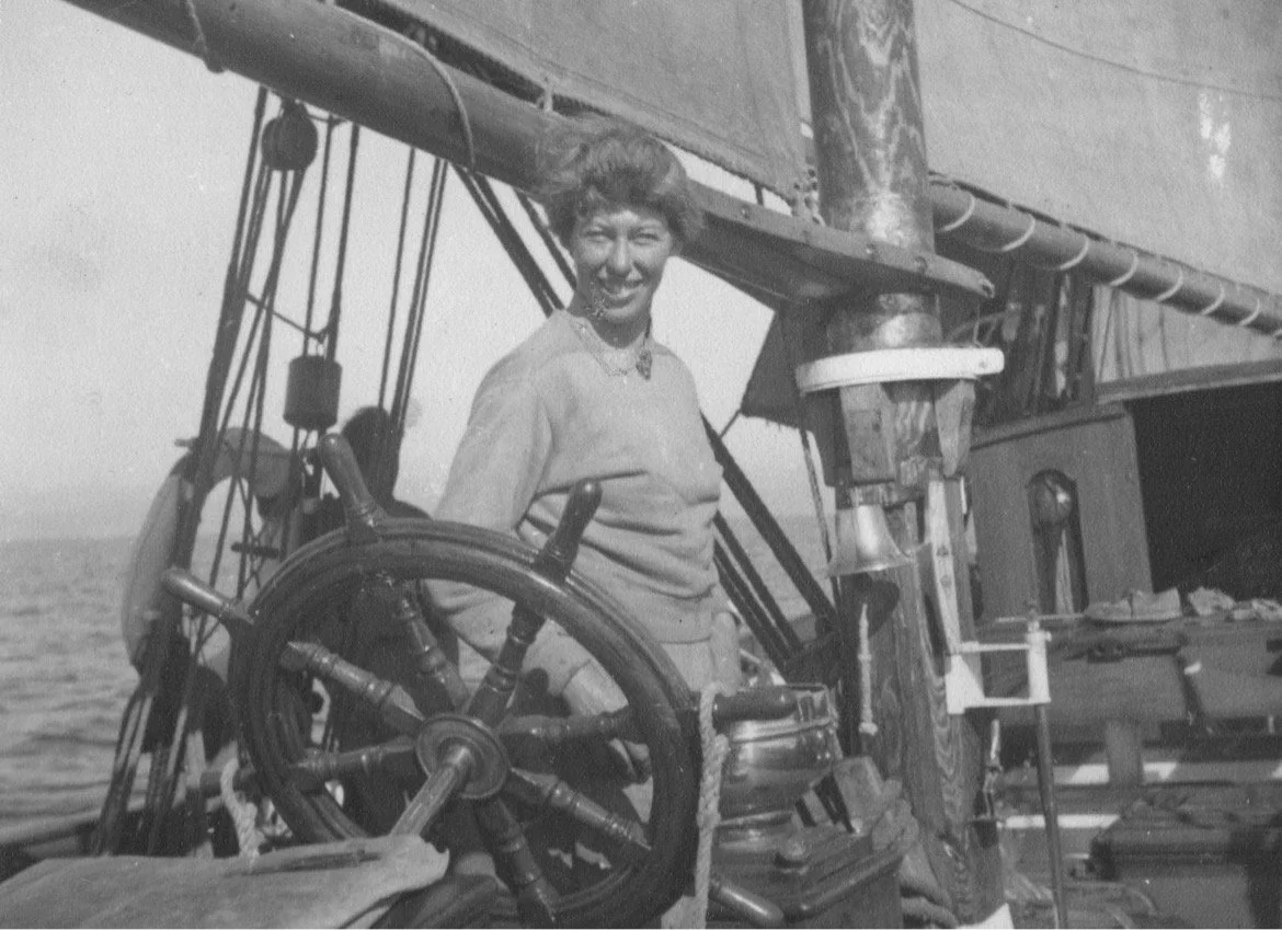 Black and white photo of a woman standing on Provident, smiling, with a large ship's wheel in front of her.