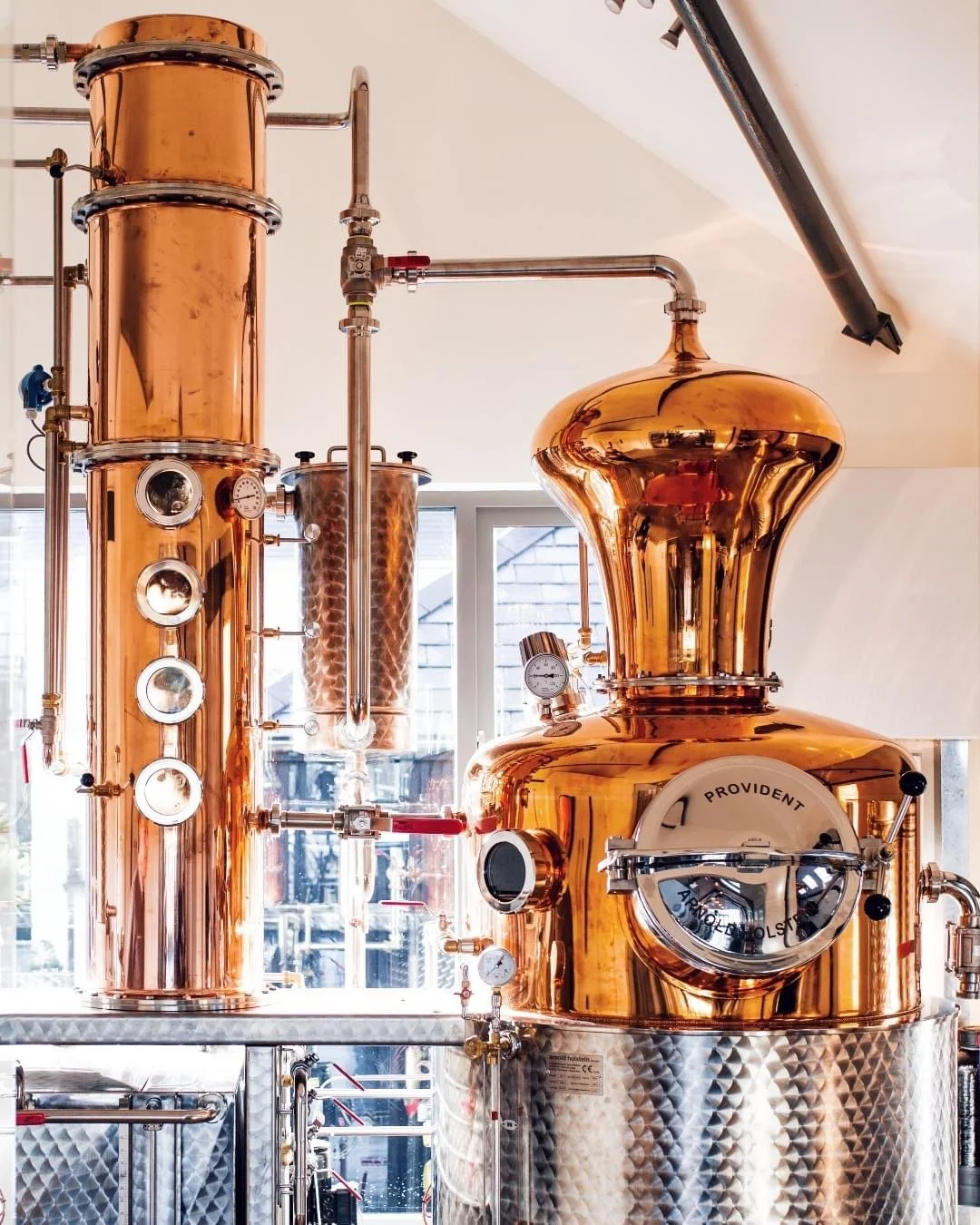 Copper distillation apparatus with gauges and pipes in a bright room, likely used for alcohol production or similar process.
