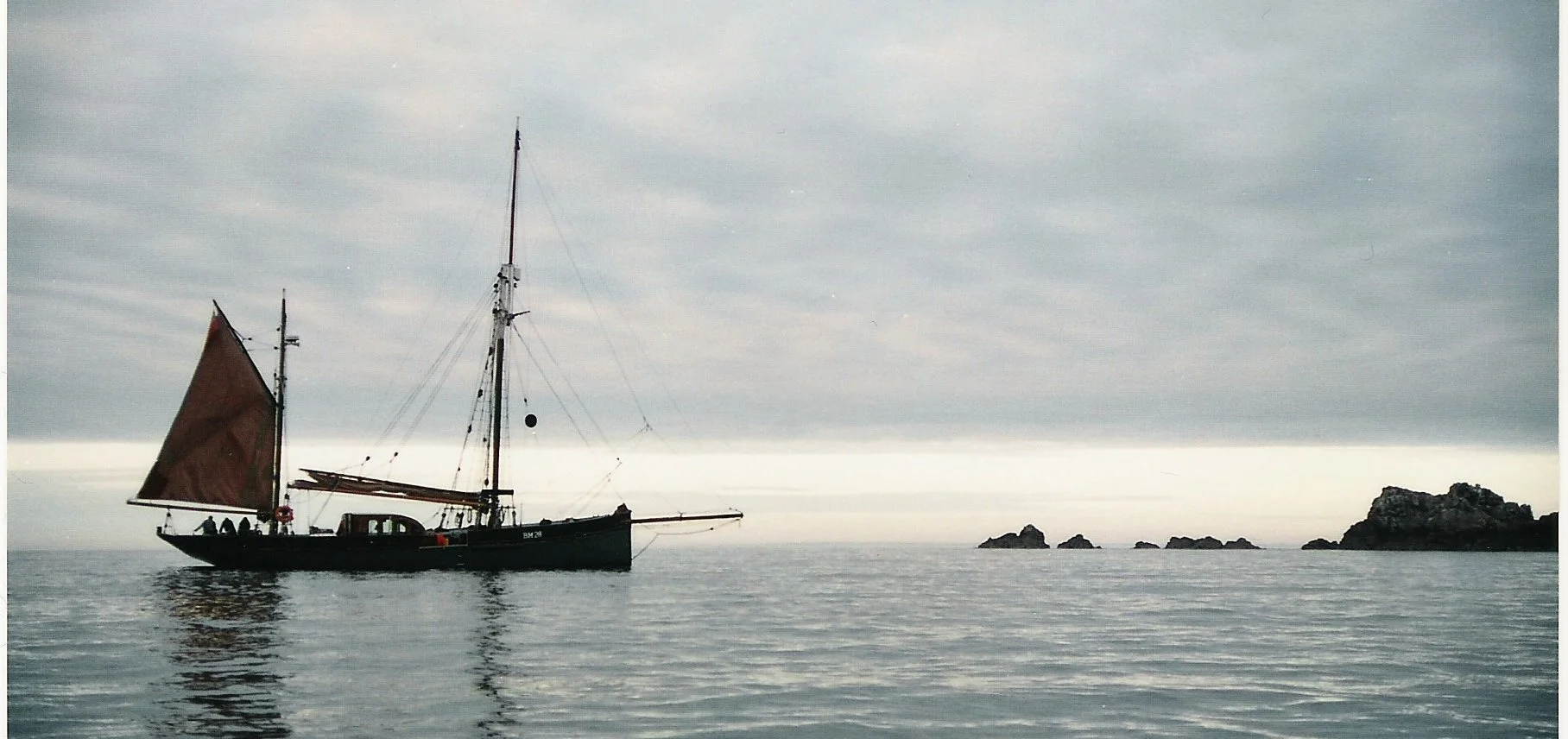 Provident with a brown sail and a black hull floating on calm water with rocky islands in the distance under an overcast sky.