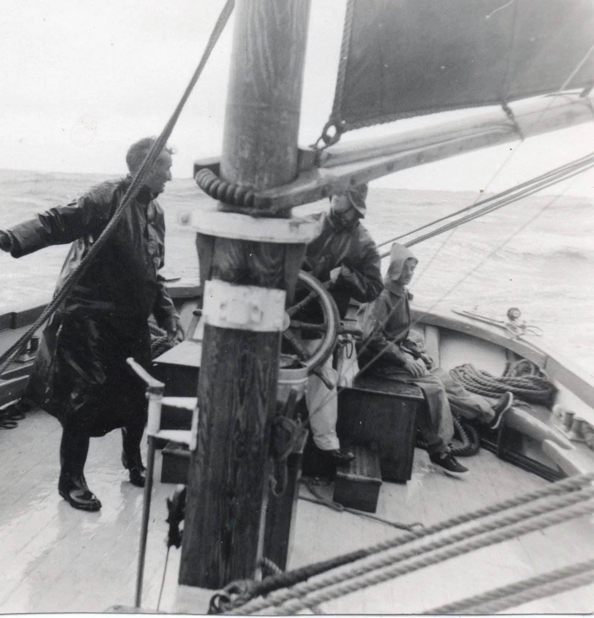 Black and white photo of three people on Provident, with one person standing and two sitting, near the ship's helm, on a body of water with an overcast sky.