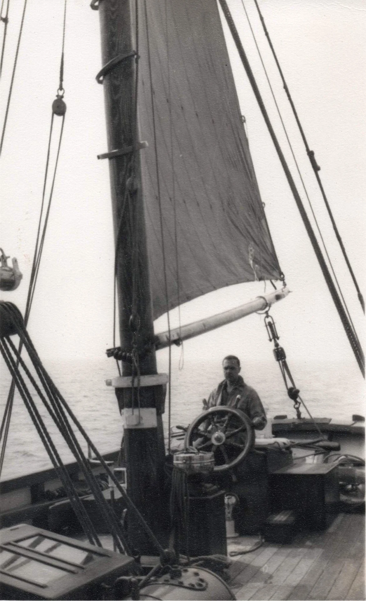 Black and white photo of a man standing at the helm of Provident, with the sea in the background. The sailboat's mast and rigging are visible.