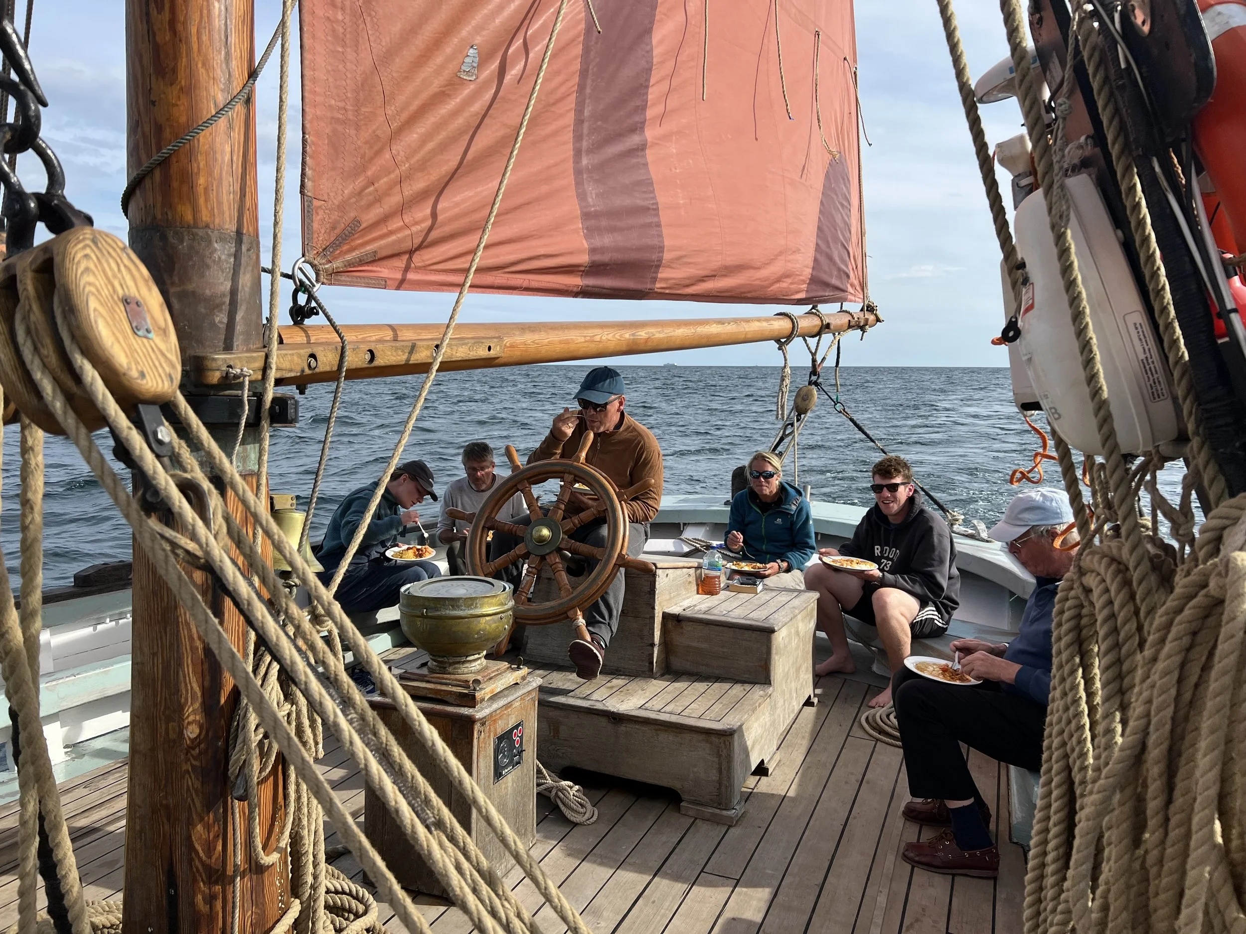 Group of people enjoying a meal on Provident with an orange sail, surrounded by ropes, on the water during the day.