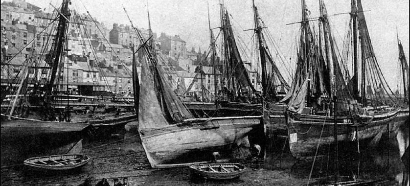 Old black-and-white photograph of several sailboats docked at a harbor with buildings on a hillside in the background.