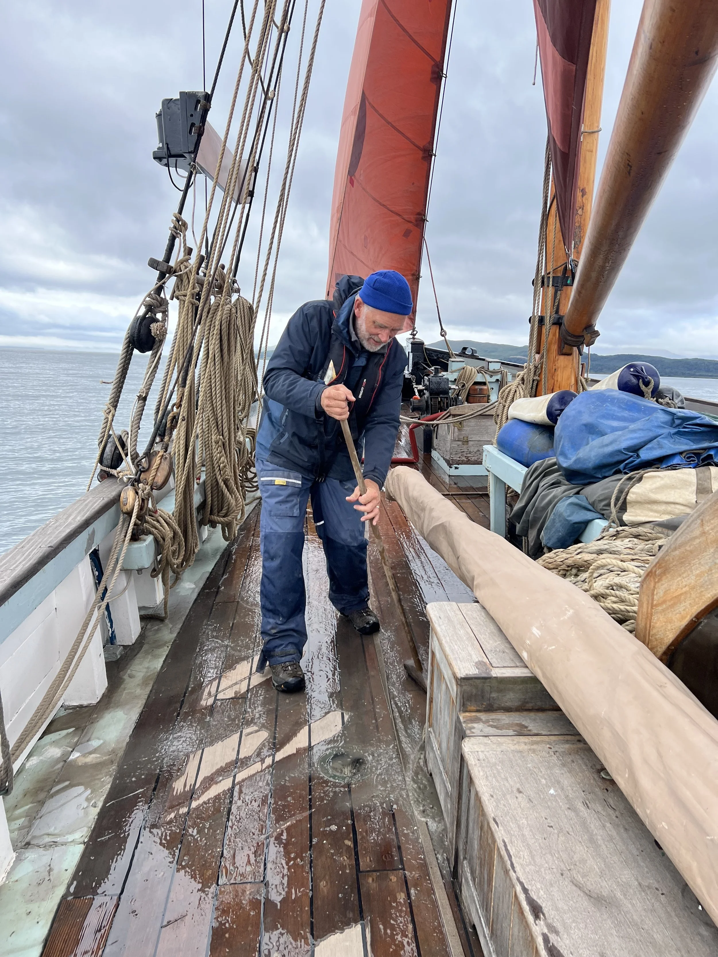 A man in waterproof clothing sweeping the wet wooden deck of Provident with a broom, on a cloudy day at sea.