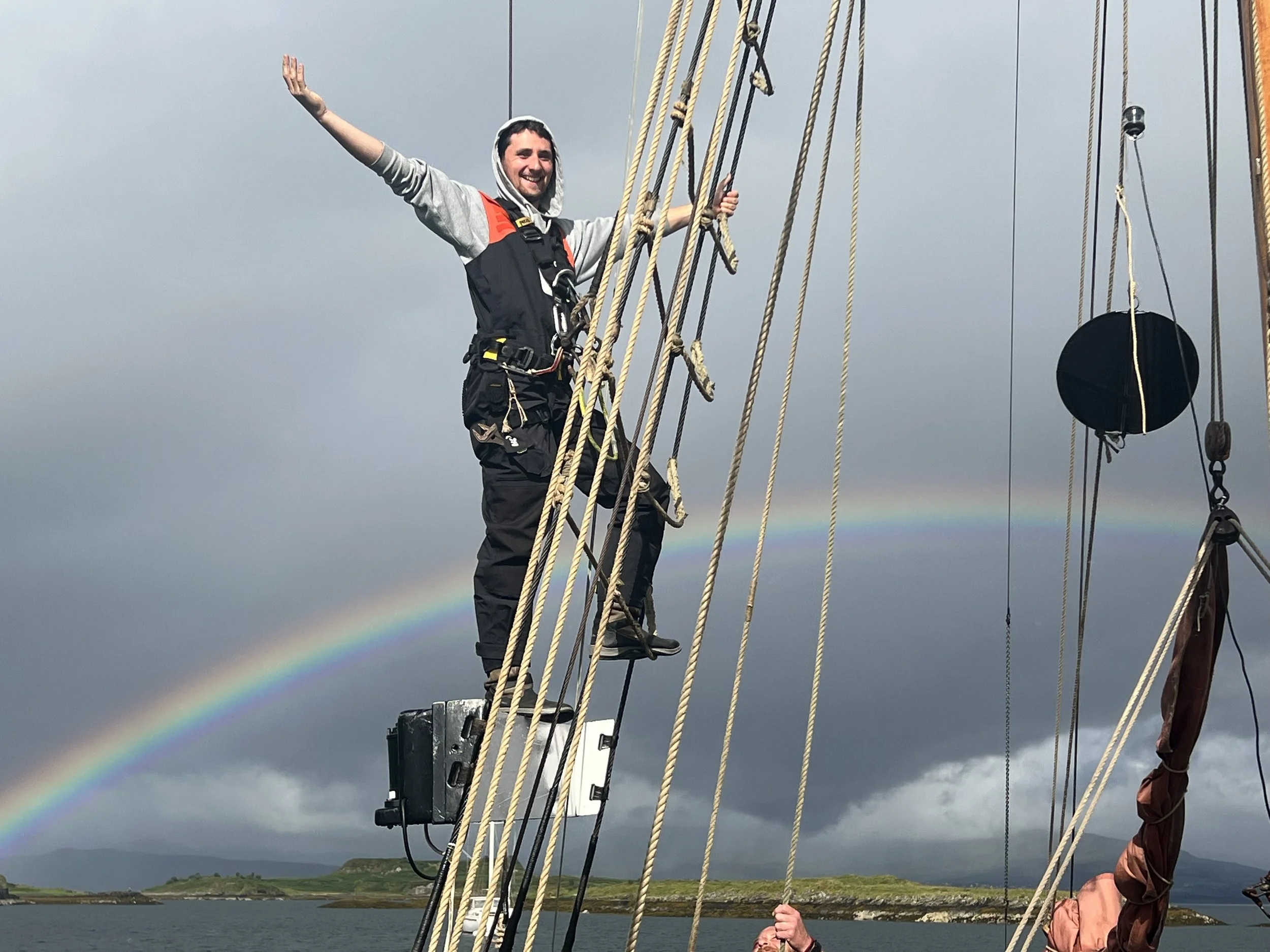A man standing on the mast of Provident, smiling and waving with a rainbow in the cloudy sky behind him.