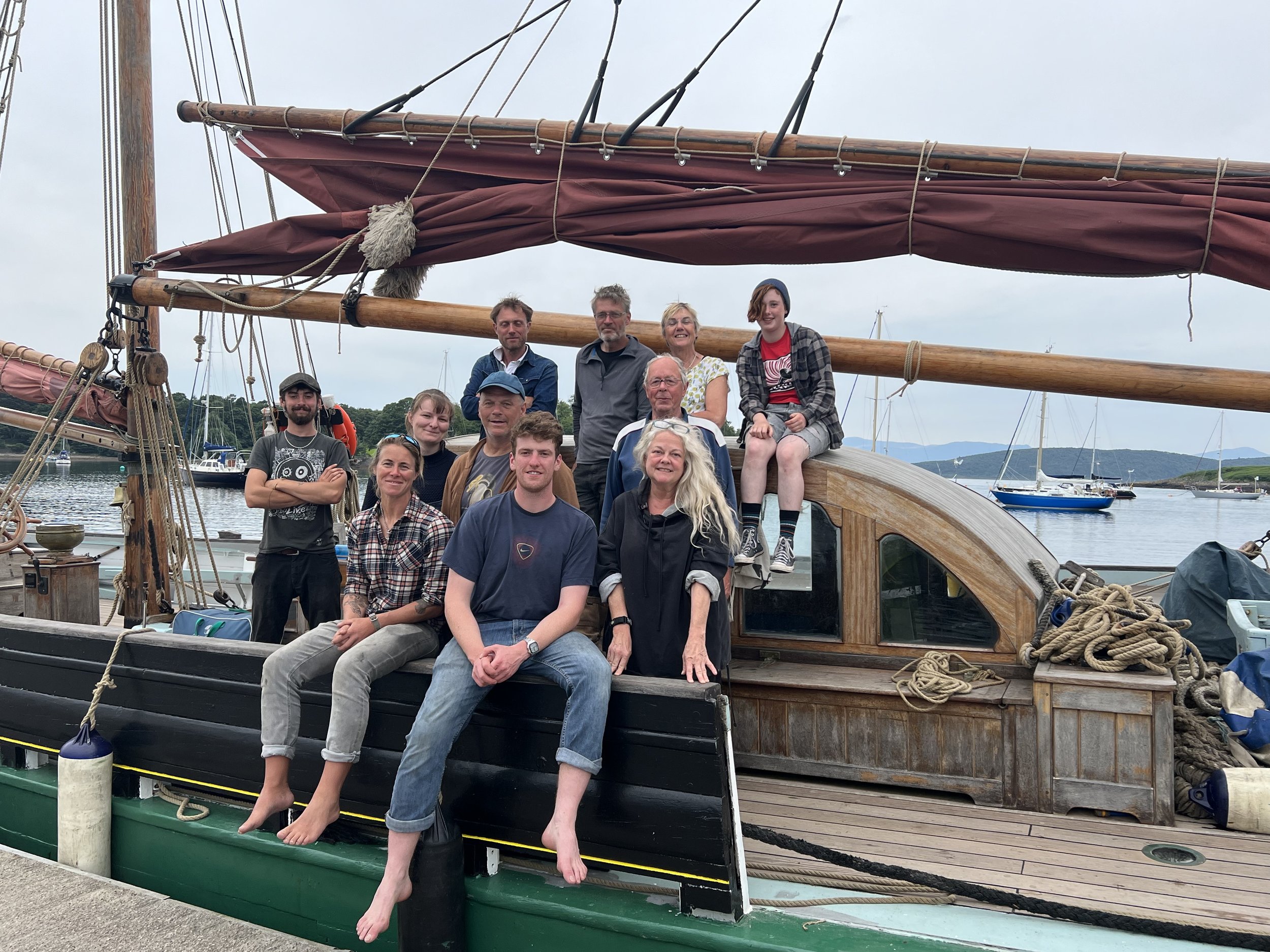 Group of people posing on Provident with a maroon sail, with water and other sailboats in the background.