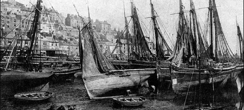 Black and white photo of multiple sailing ships docked at a harbor with buildings on a hillside in the background.