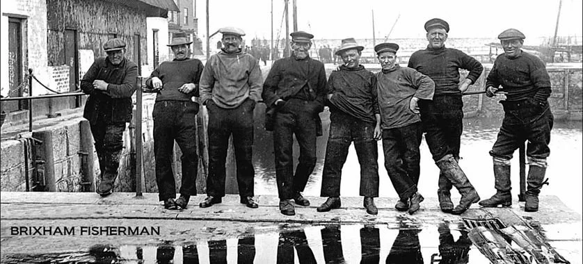 Historical black-and-white photo of eight fishermen standing on a dock, with boats in the background, by the water. The text 'BRIXHAM FISHERMAN' is visible in the bottom left corner.