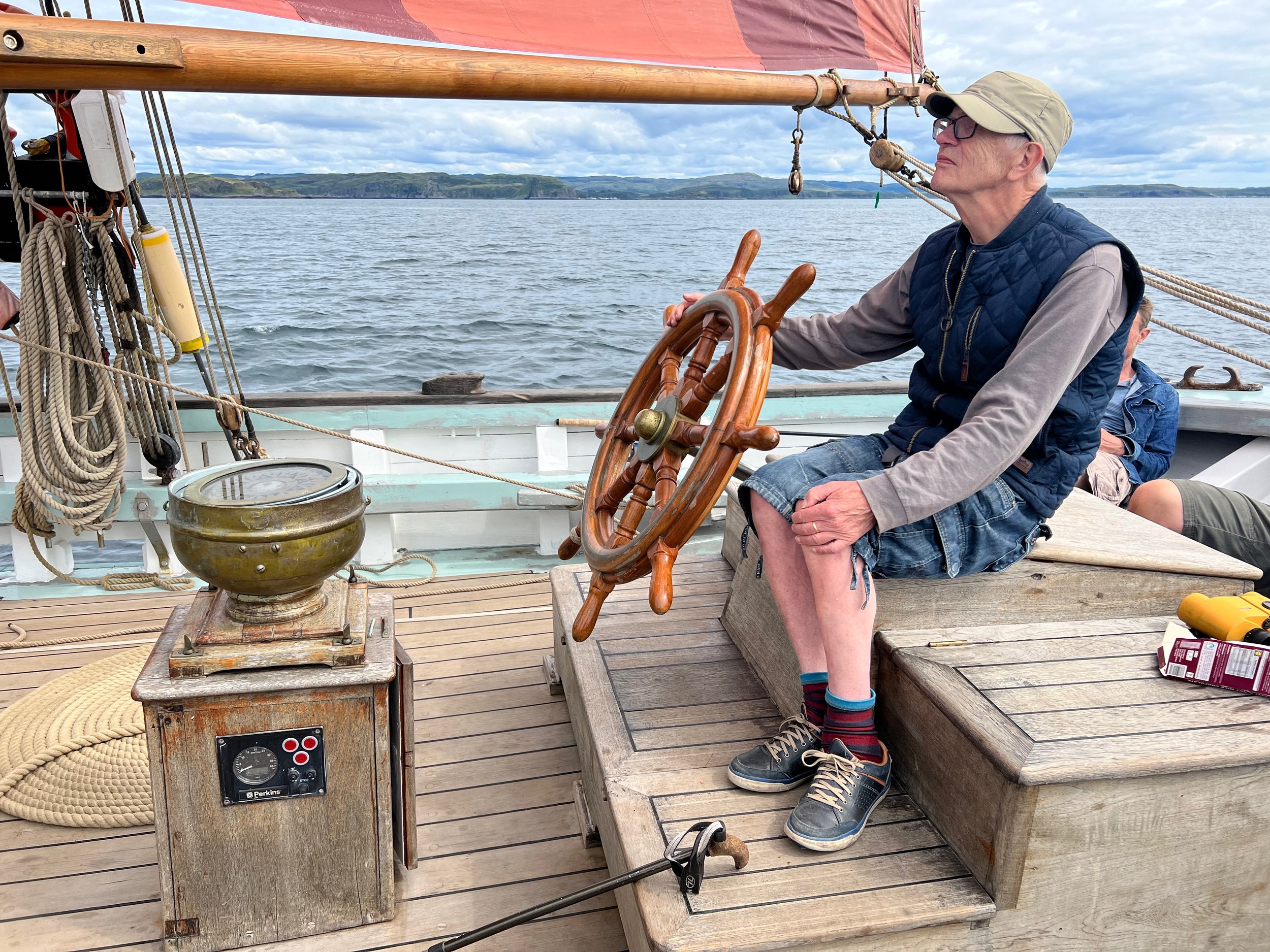 A man sitting on Provident's deck holding Provident's wheel, wearing a cap, glasses, and casual clothing with a vest and shorts, with water and distant landscape in the background.