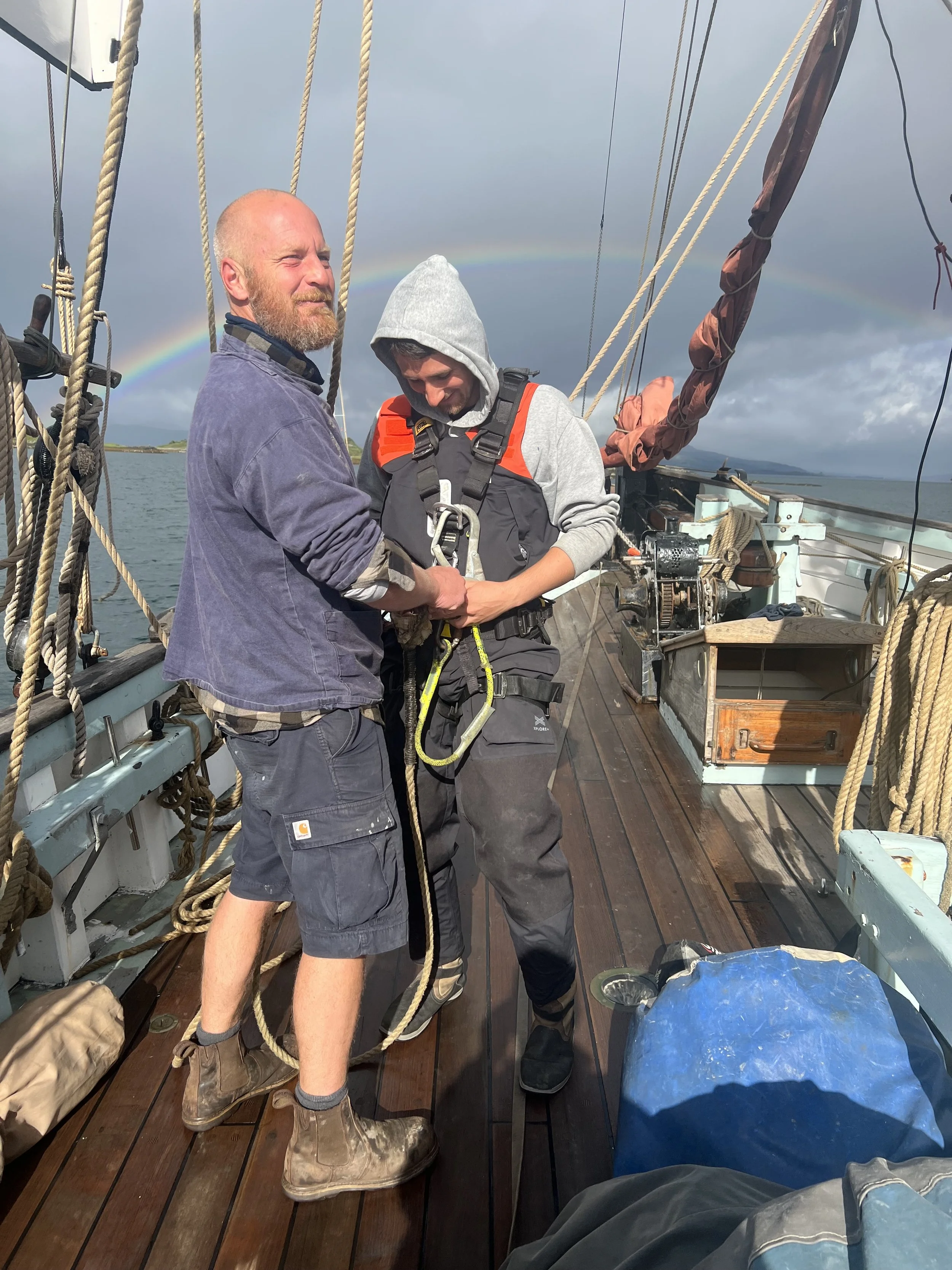 Two men on Provident's deck, one in a blue jacket and the other in a gray hoodie and harness, with a rainbow in the background.