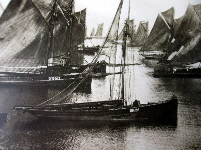 Black and white photograph of a harbor with several sailing ships, some with sails up, docked side by side.