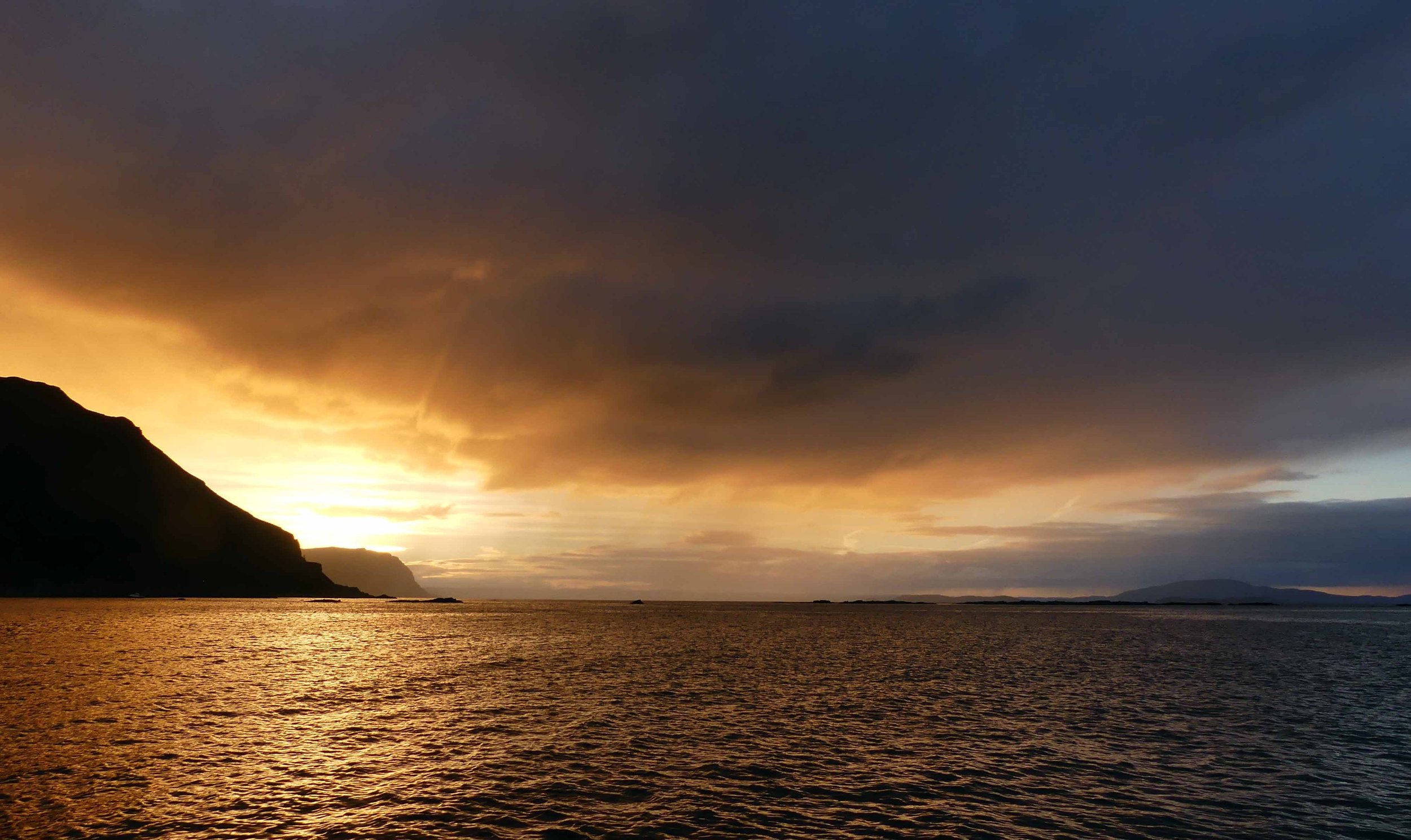 Sunset over a body of water with a silhouette of a mountain on the left and dark clouds overhead.