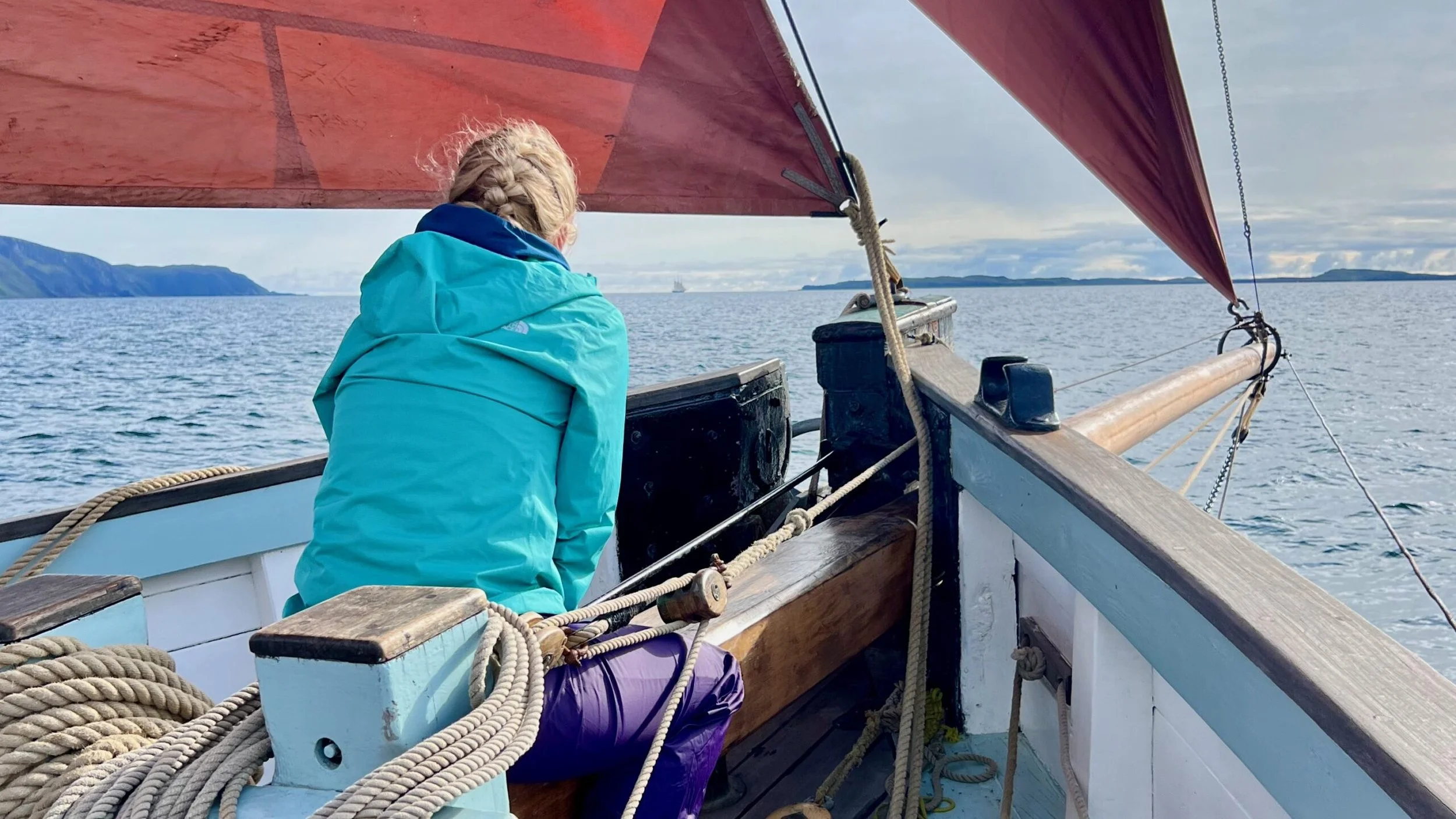 Personal  Provident looking out at the water, with land in the distance, wearing a blue jacket and braided blonde hair, under a red sail.