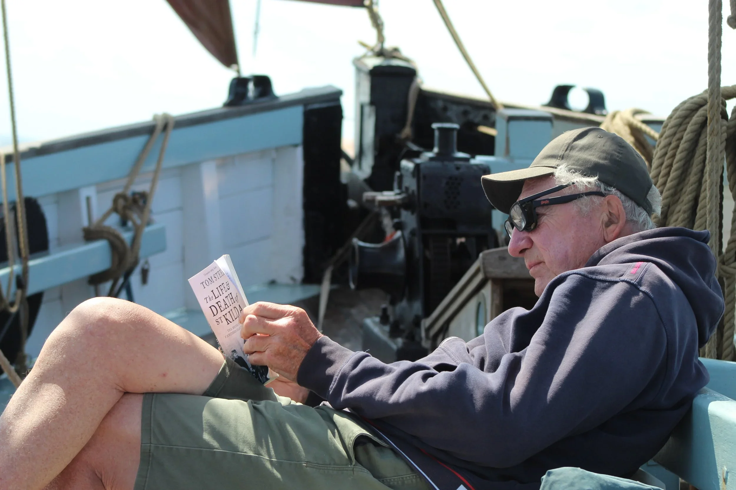 An elderly man with a gray cap, sunglasses, and a gray hoodie is sitting on Provident, reading a book titled 'The Life & Death of St. Kilda' by Tom Steel.