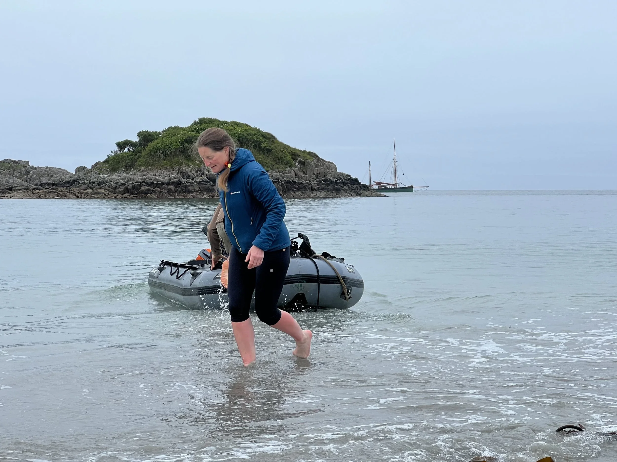 Woman in a blue jacket stepping out of the water near a boat with a small island and Provident  in the background.