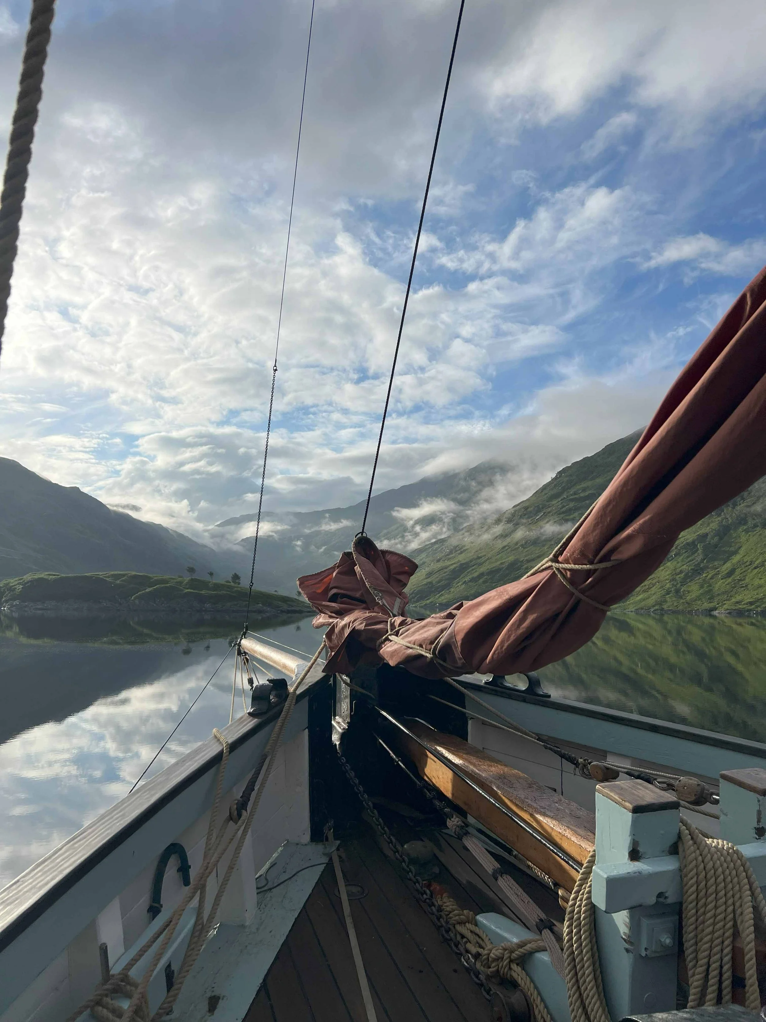 View from a boat sailing on a calm lake surrounded by green mountains with clouds and blue sky overhead.