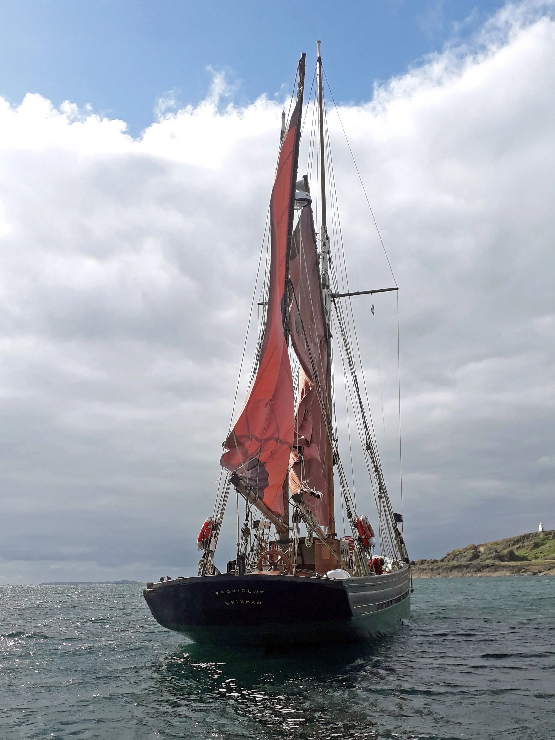 Provident with red sails on the water near a coastline with a lighthouse in the background.