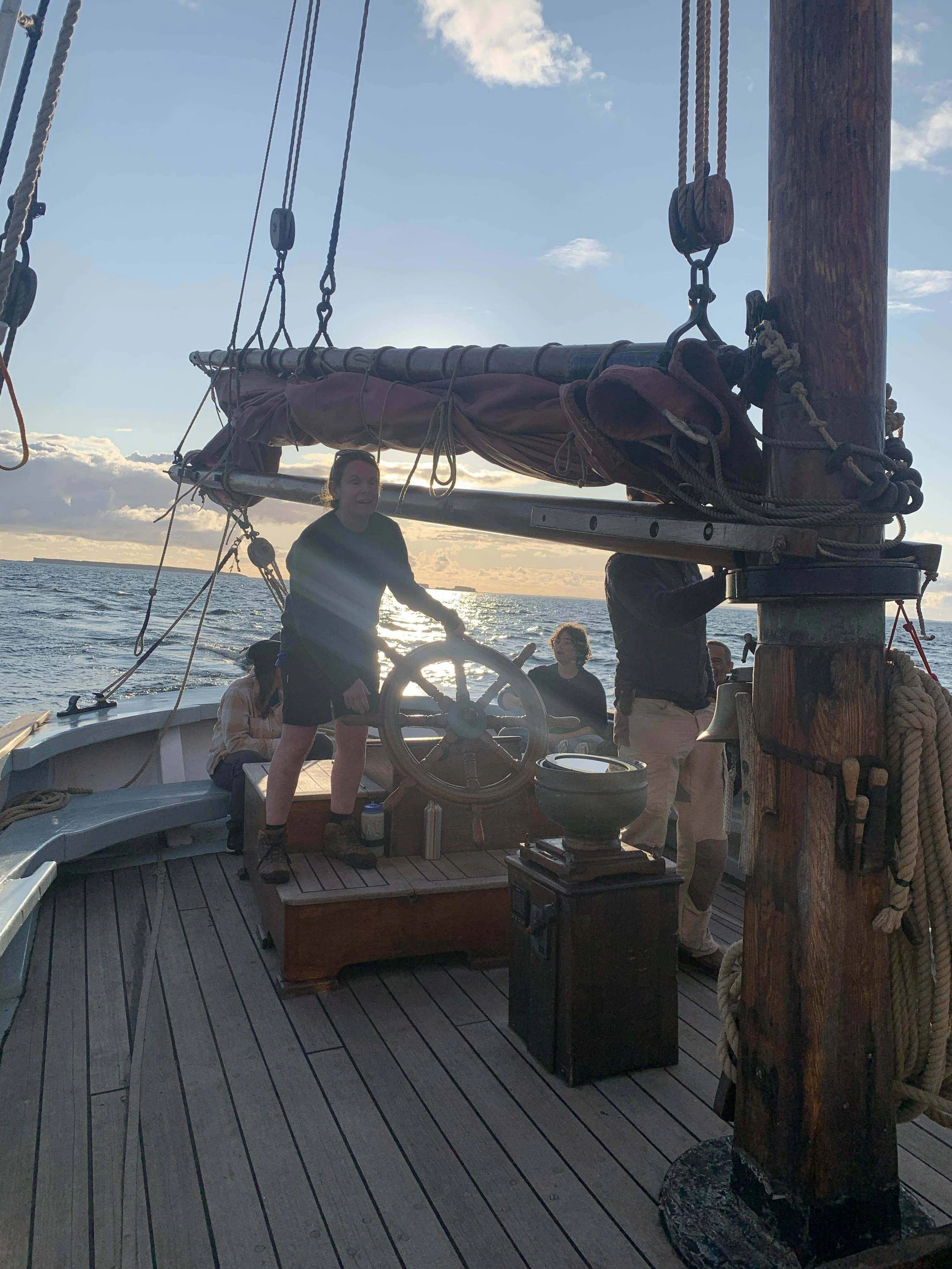 People on a Provident at sunset, one person standing at the helm steering, others sitting and standing nearby, ocean and sky with clouds in the background.