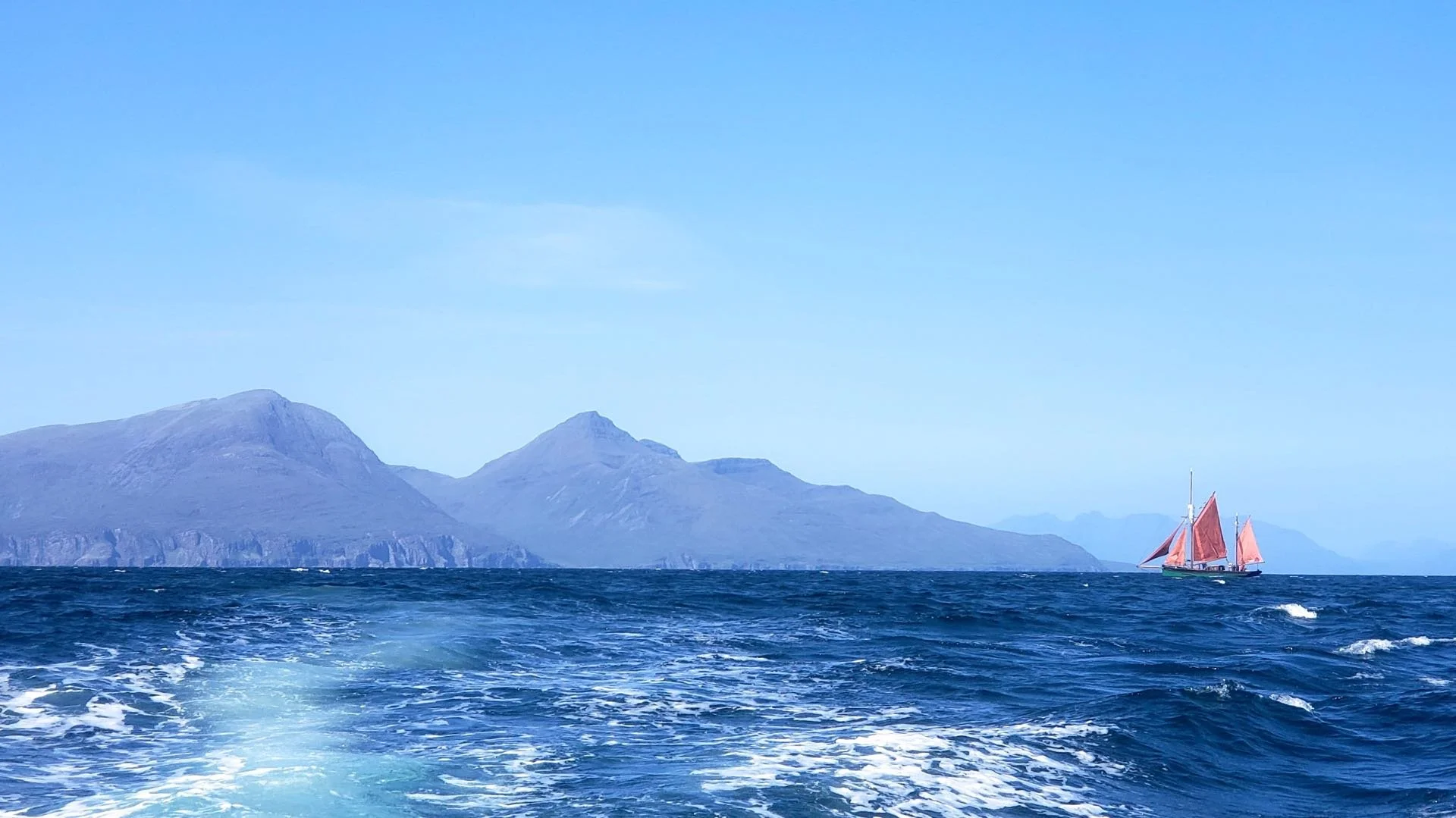 The image shows a body of water with Provident in the distance, gold with red sails, and a mountain range in the background under a clear blue sky.