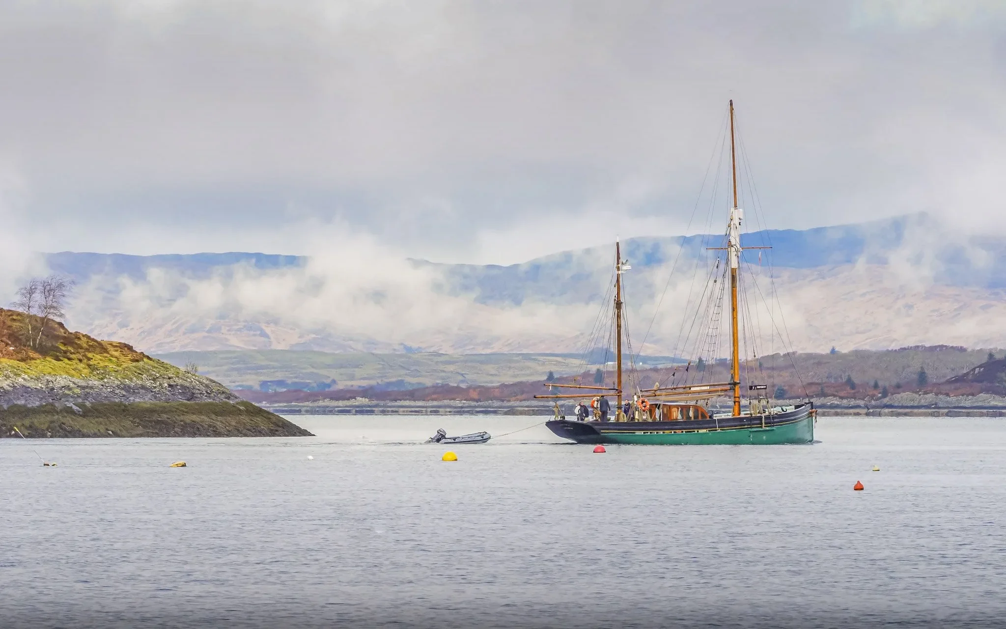 Provident with two masts and a small dinghy attached, floating on calm water near a hilly coastline with distant mountains.