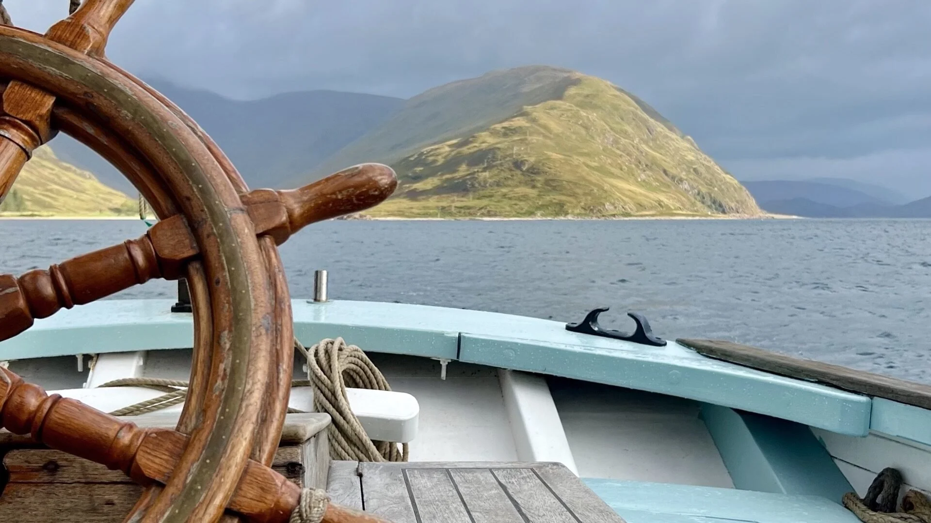 View from a boat showing a wooden steering wheel, ropes, and part of the boat's deck with a mountain in the background under a cloudy sky.