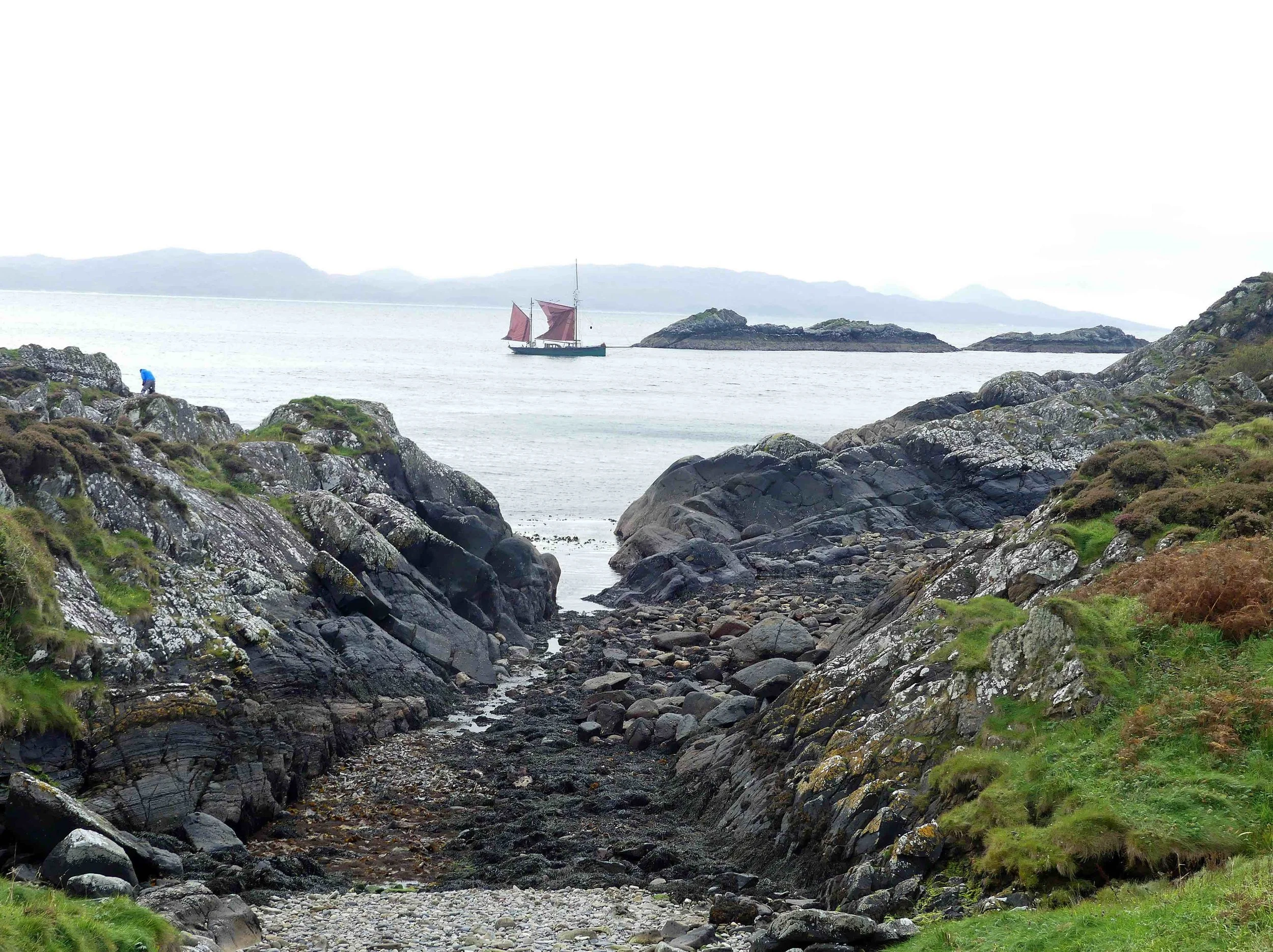 A rocky coastal scene with a narrow, rocky pathway leading to the sea. A person in a blue jacket is standing on the rocks to the left, and a sailboat with red sails is in the water. Distant landforms are visible on the horizon under a cloudy sky.