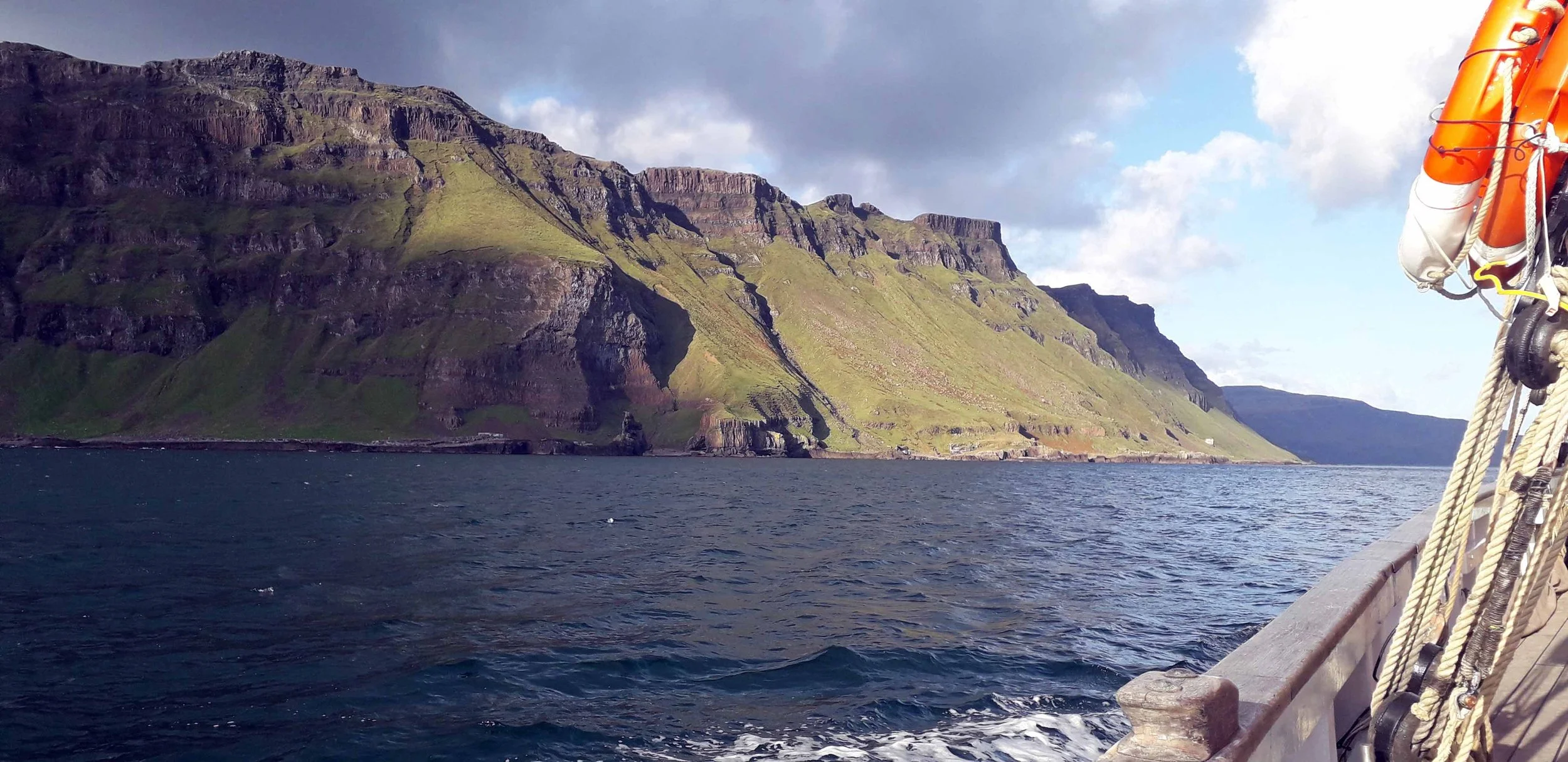View from a ship showing a large green, rocky island with steep cliffs and a body of water in the foreground. Part of the ship's deck and ropes are visible on the right side.