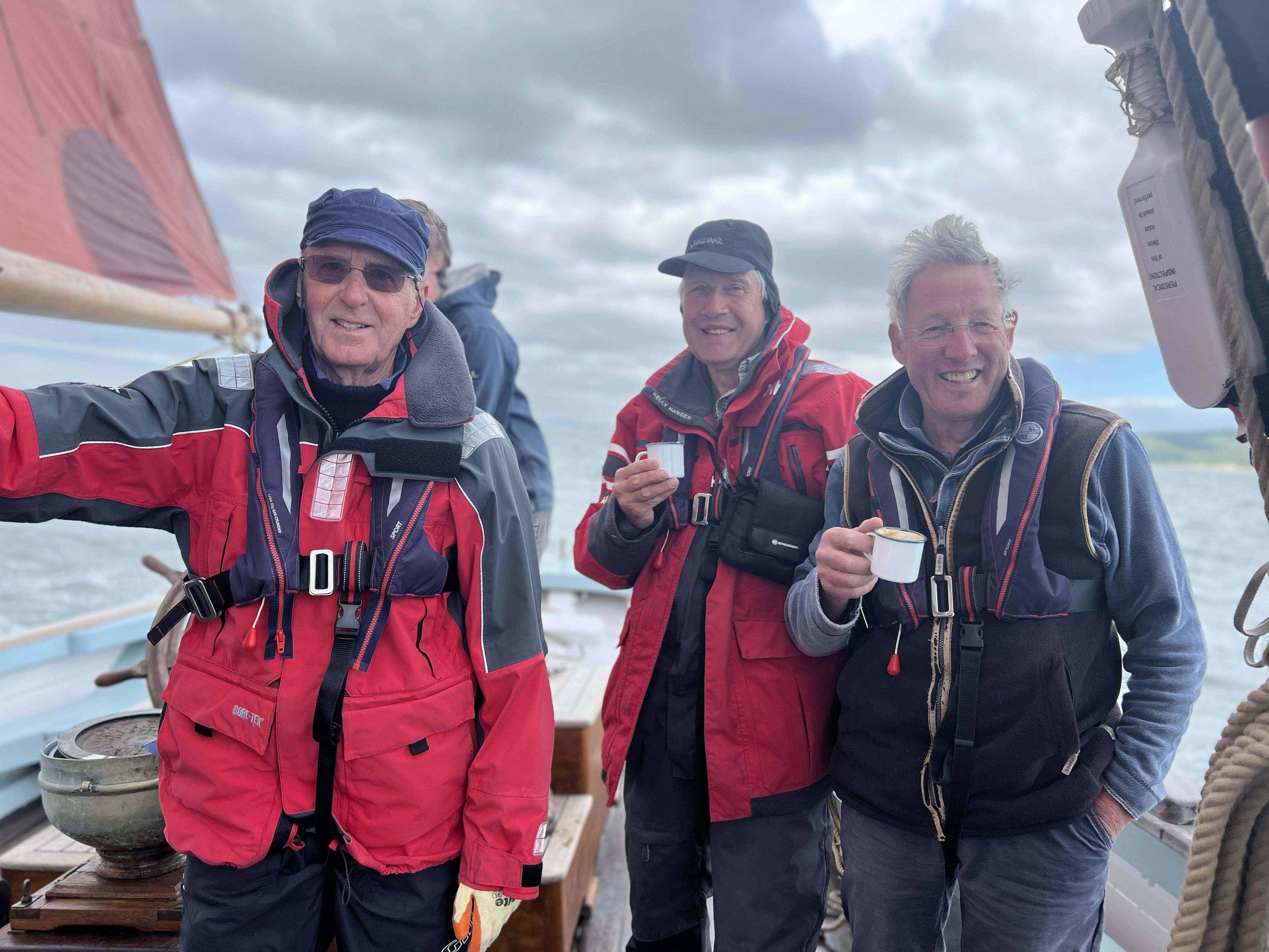 Three older men on Provident wearing outdoor jackets, holding small cups, smiling, with gray clouds in the sky and water in the background.