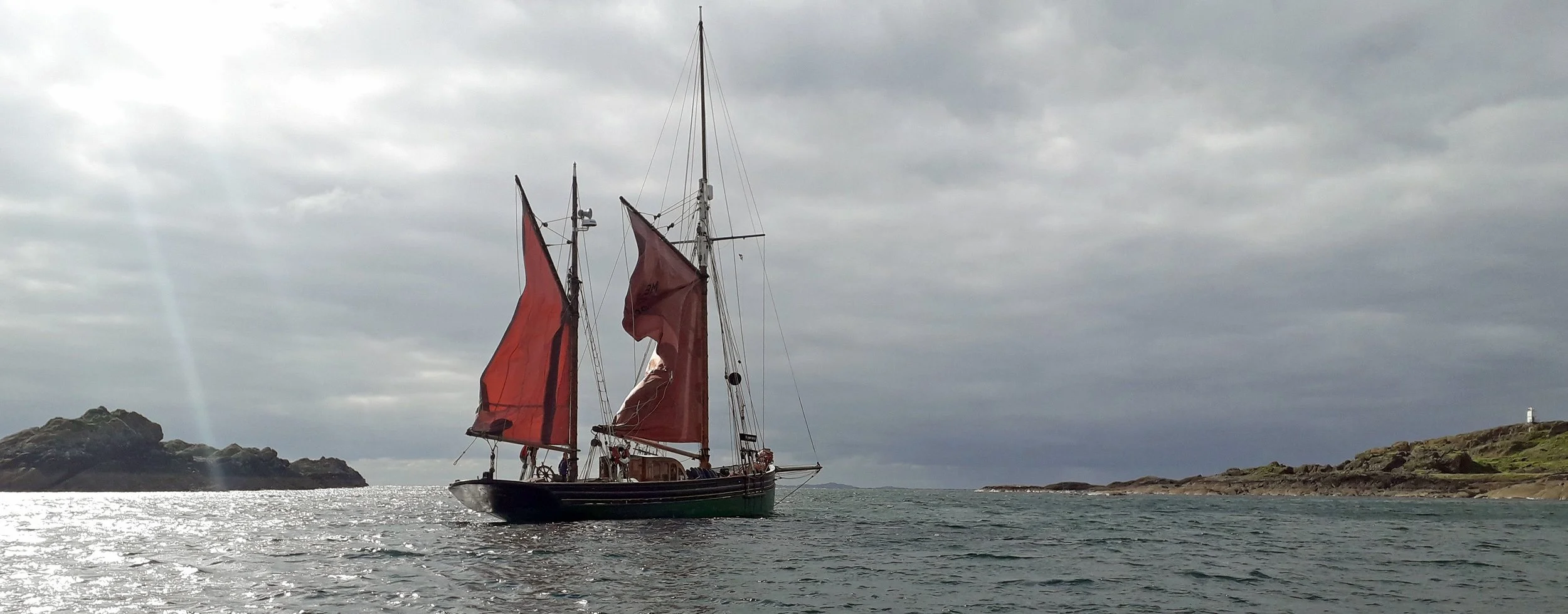 Provident with red sails sailing on a calm ocean near rocky land under a cloudy sky.