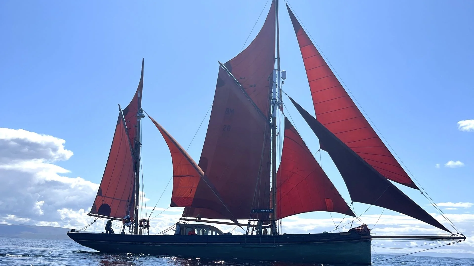 Provident with large red and black sails sailing on calm water against a blue sky with some clouds.