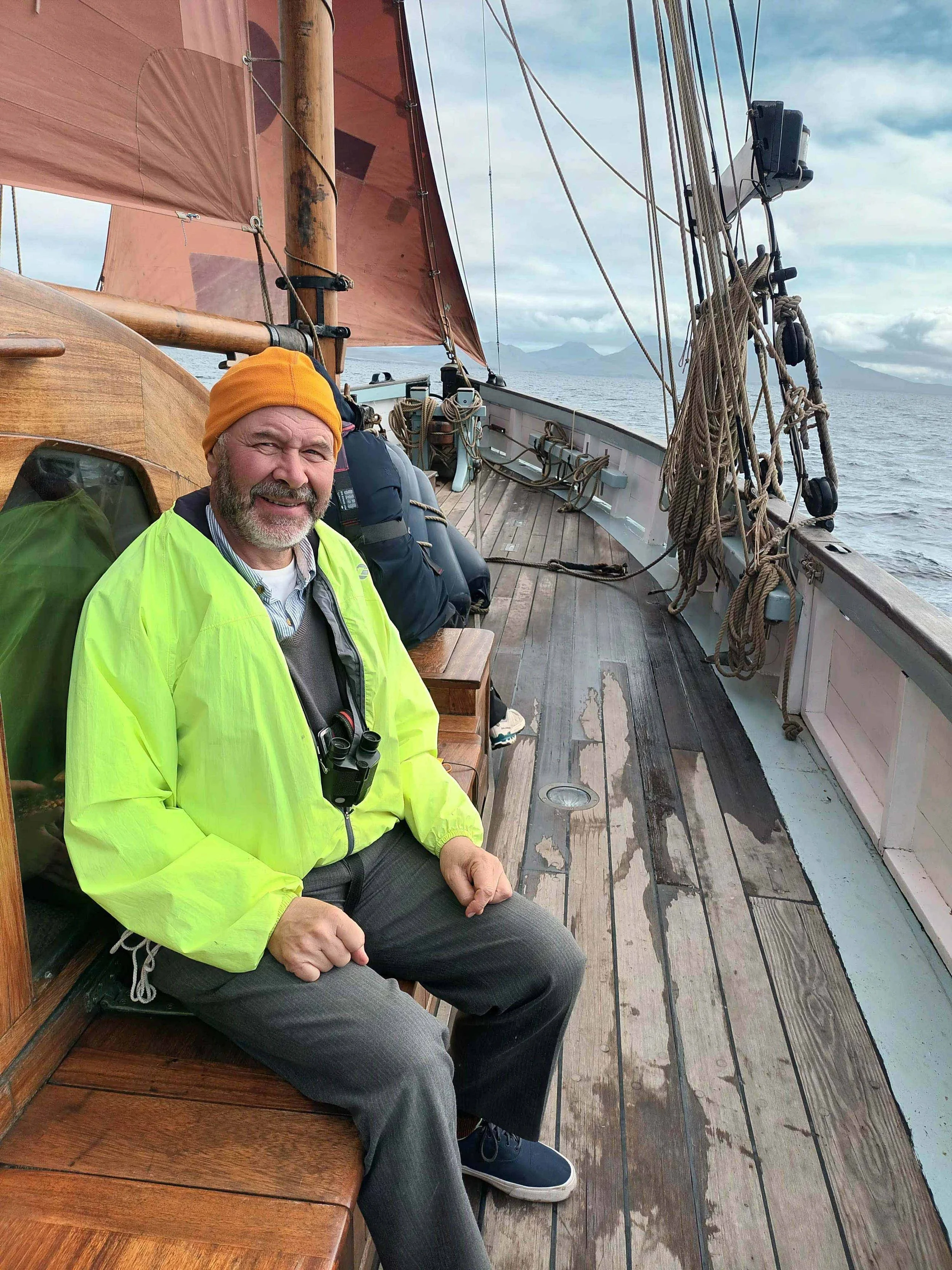 A man with gray hair and a beard sitting on a wooden boat deck, wearing a yellow jacket, orange beanie, and gray pants, with binoculars hanging around his neck. The boat is on the water with mountains and cloudy sky in the background.
