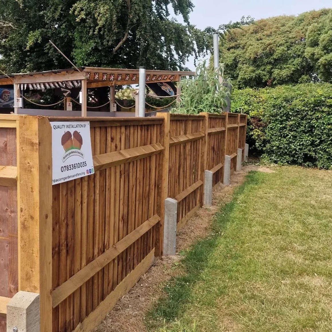 A wooden fence with support posts along a grassy area, with trees and shrubs in the background. There is a sign on the fence advertising a gardening and fencing installation service.