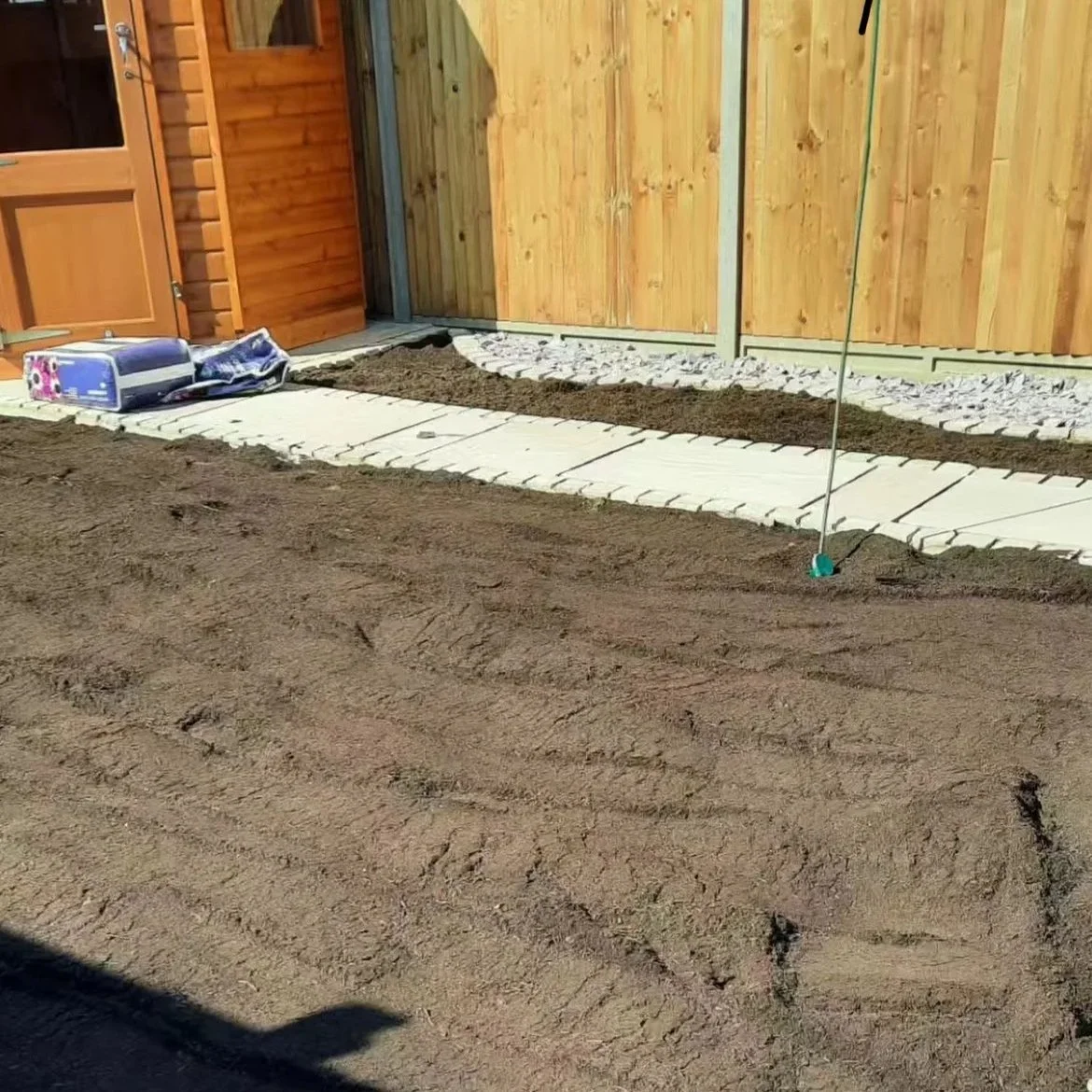 A backyard area under construction with a path made of light-colored paving stones on a bed of soil, next to a wooden fence and a wooden shed.