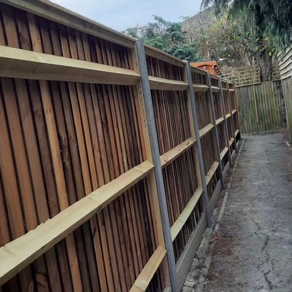 Fence panels leaning against a metal support frame along a narrow outdoor pathway, with trees and a wooden fence in the background.