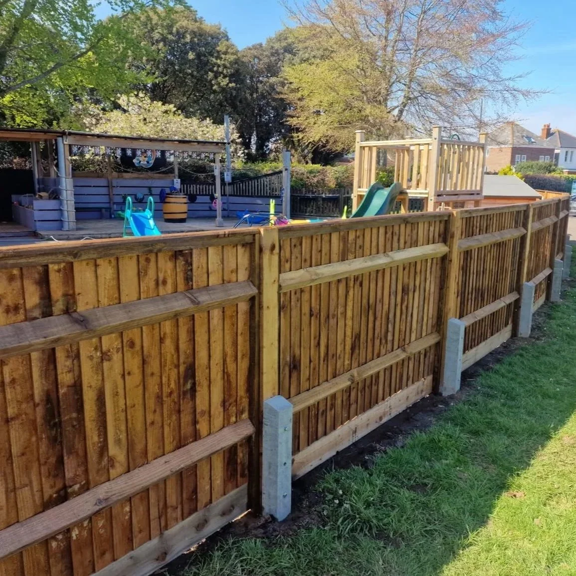A wooden fence surrounding a backyard play area with a slide and playhouse, with trees and houses in the background.