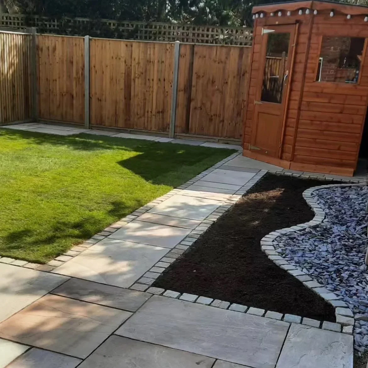 A backyard with a green lawn, a paved walkway, a wooden shed, and a wooden fence.