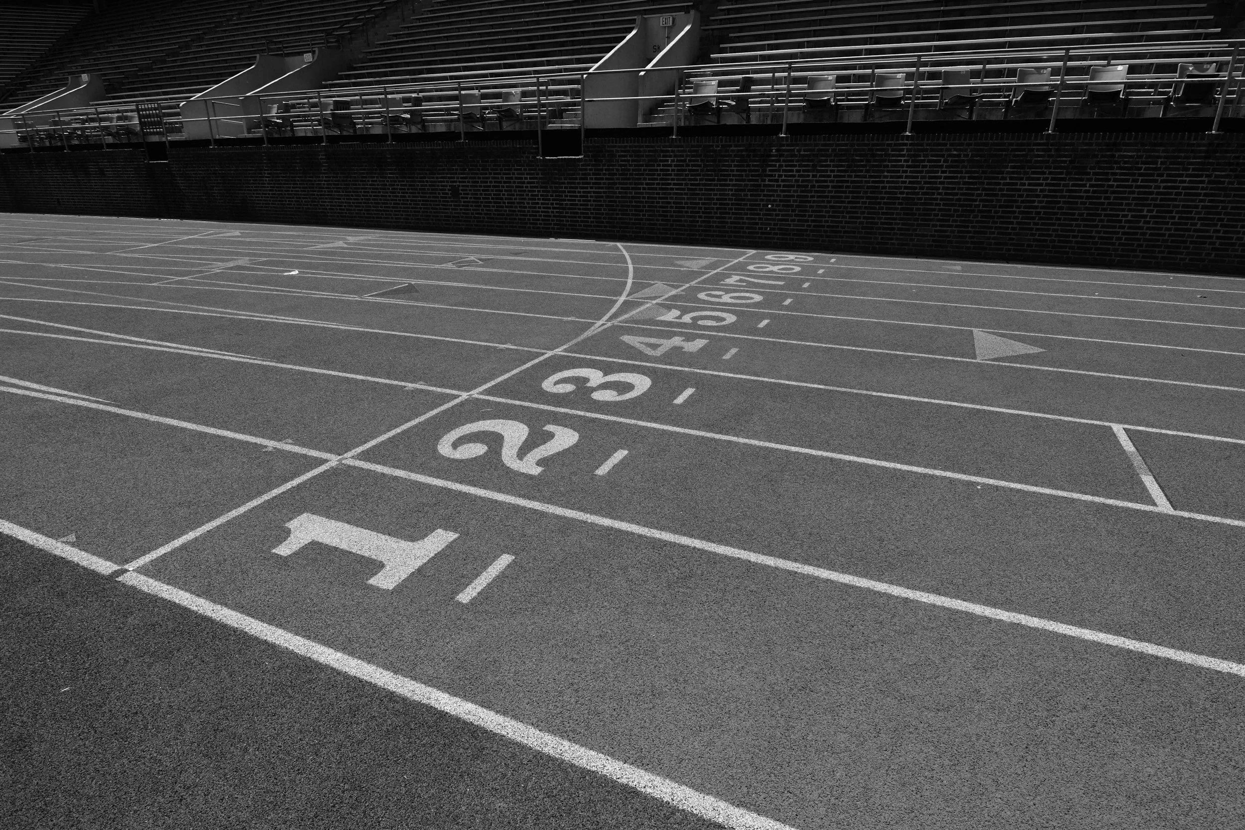 Leere Laufbahn mit nummerierten Startblöcken auf einer Leichtathletikbahn, im Hintergrund Sitzgelegenheiten und eine Zäunung.