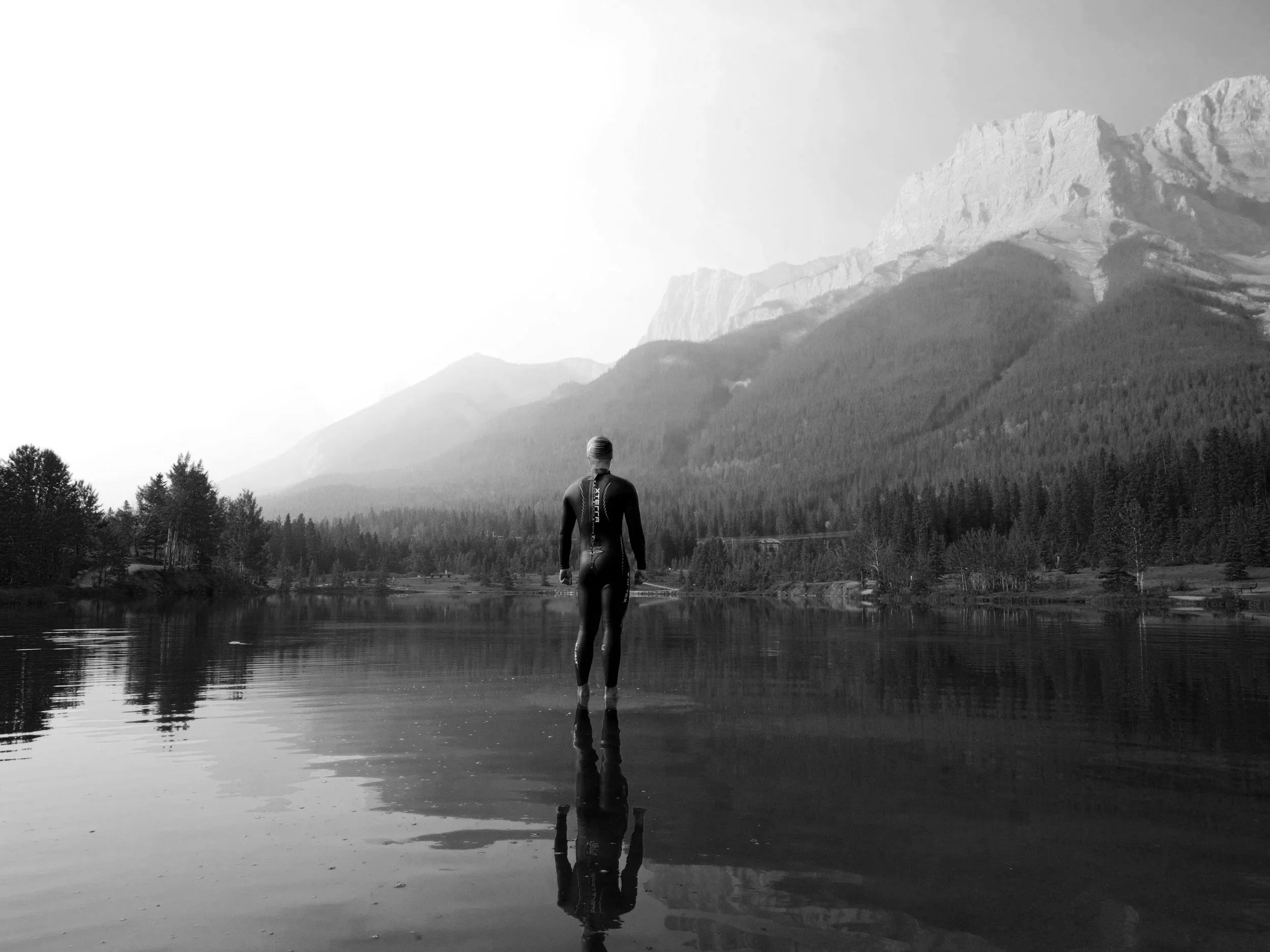 Ein Mensch in einem Neoprenanzug steht in einem See vor einer Berglandschaft, im Schwarz-Weiß-Format.