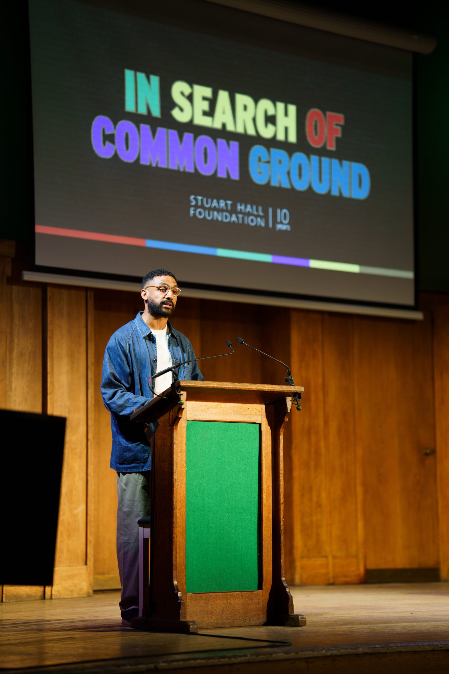 Orsod stands at a wooden podium on a stage, delivering a speech or presentation. Behind him is a large screen displaying the title 'In Search of Common Ground' with Stuart Hall Foundation written underneath the title.