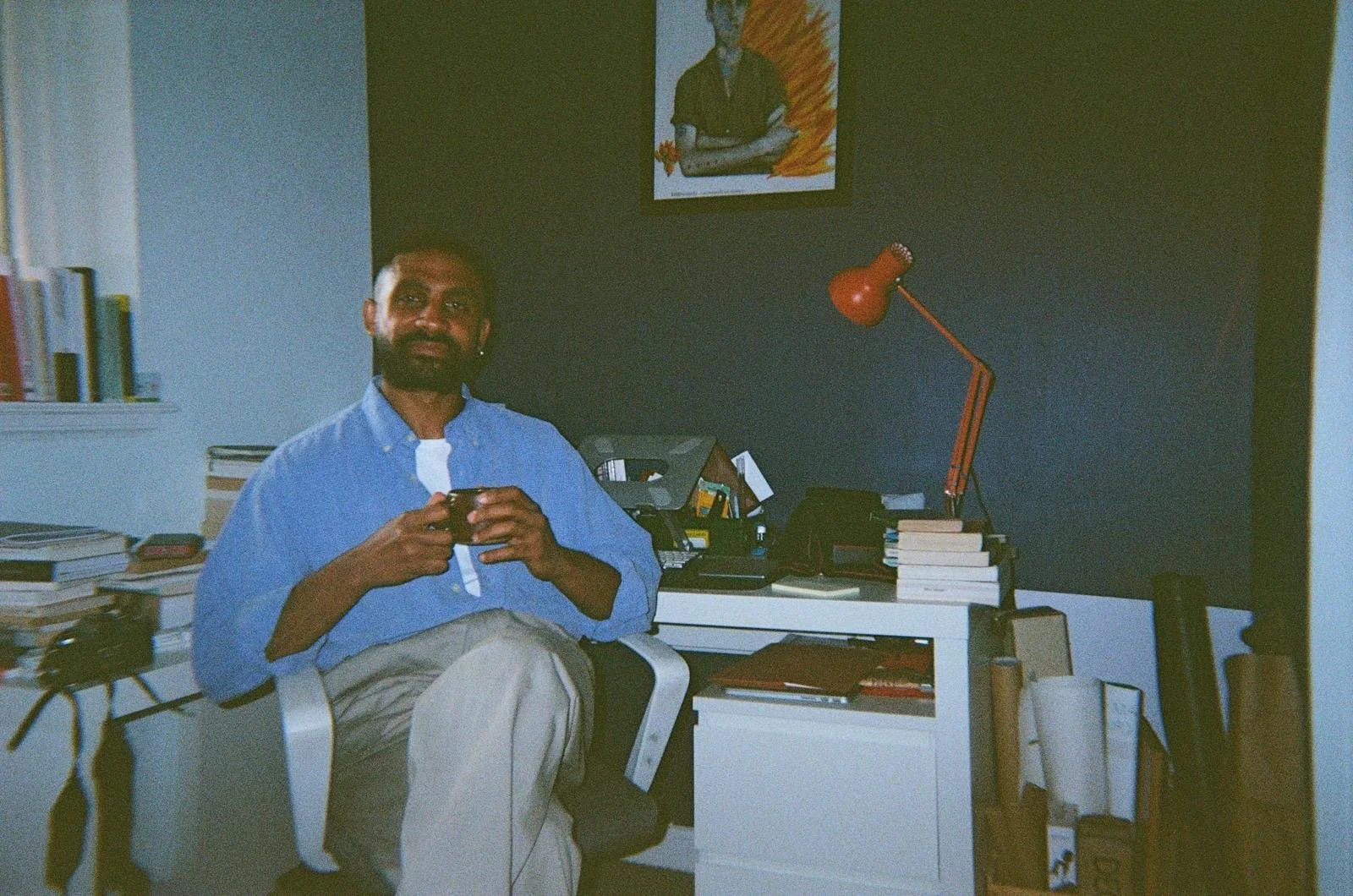 Orsod sitting in an office, holding a mug, with a desk filled with books, a red desk lamp, a telephone, and a black bag. There is a painting on the dark-colored wall behind him and a shelf with books to his left.