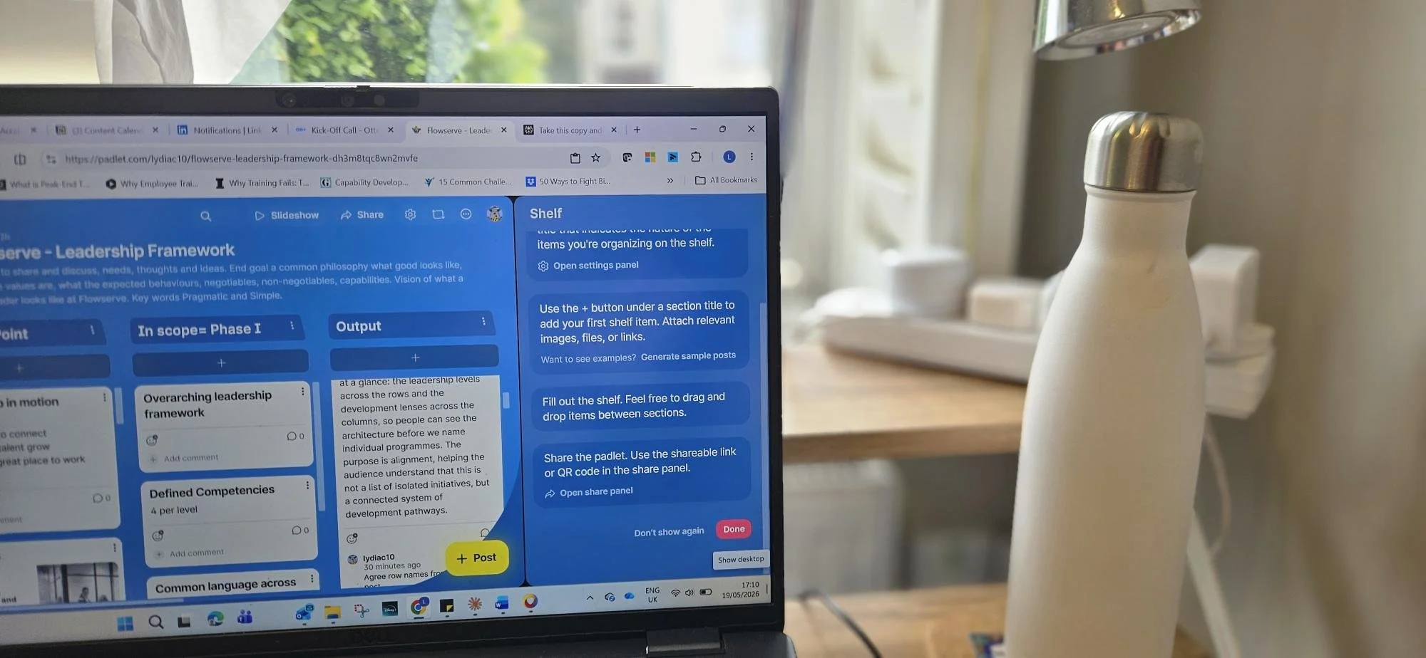 A laptop screen displaying a digital flowchart or framework, with a water bottle to the right and a blurred background of a white device and a window with natural light.