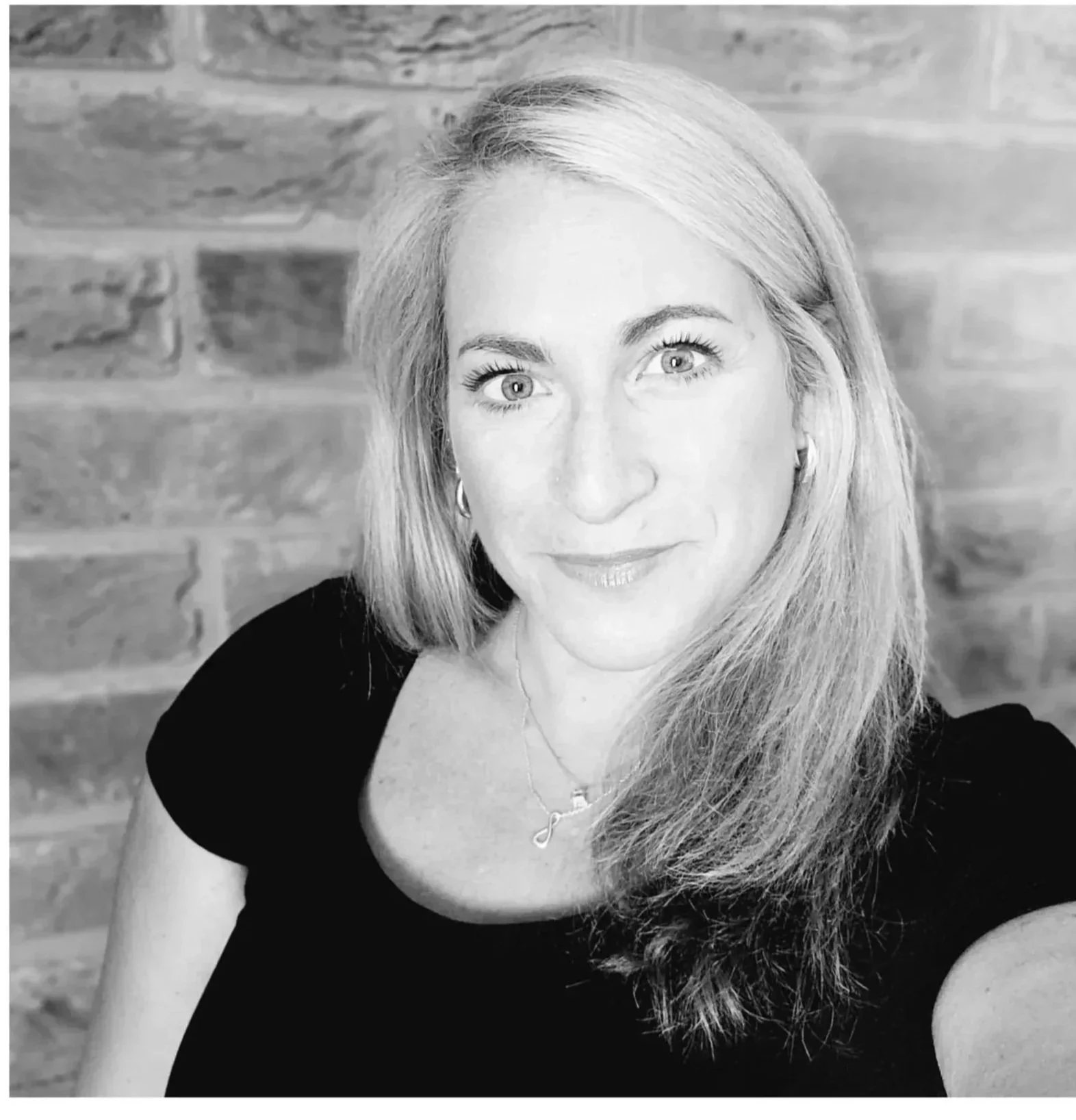 A woman with long hair smiling at the camera, wearing a black top and a necklace, with a brick wall background.