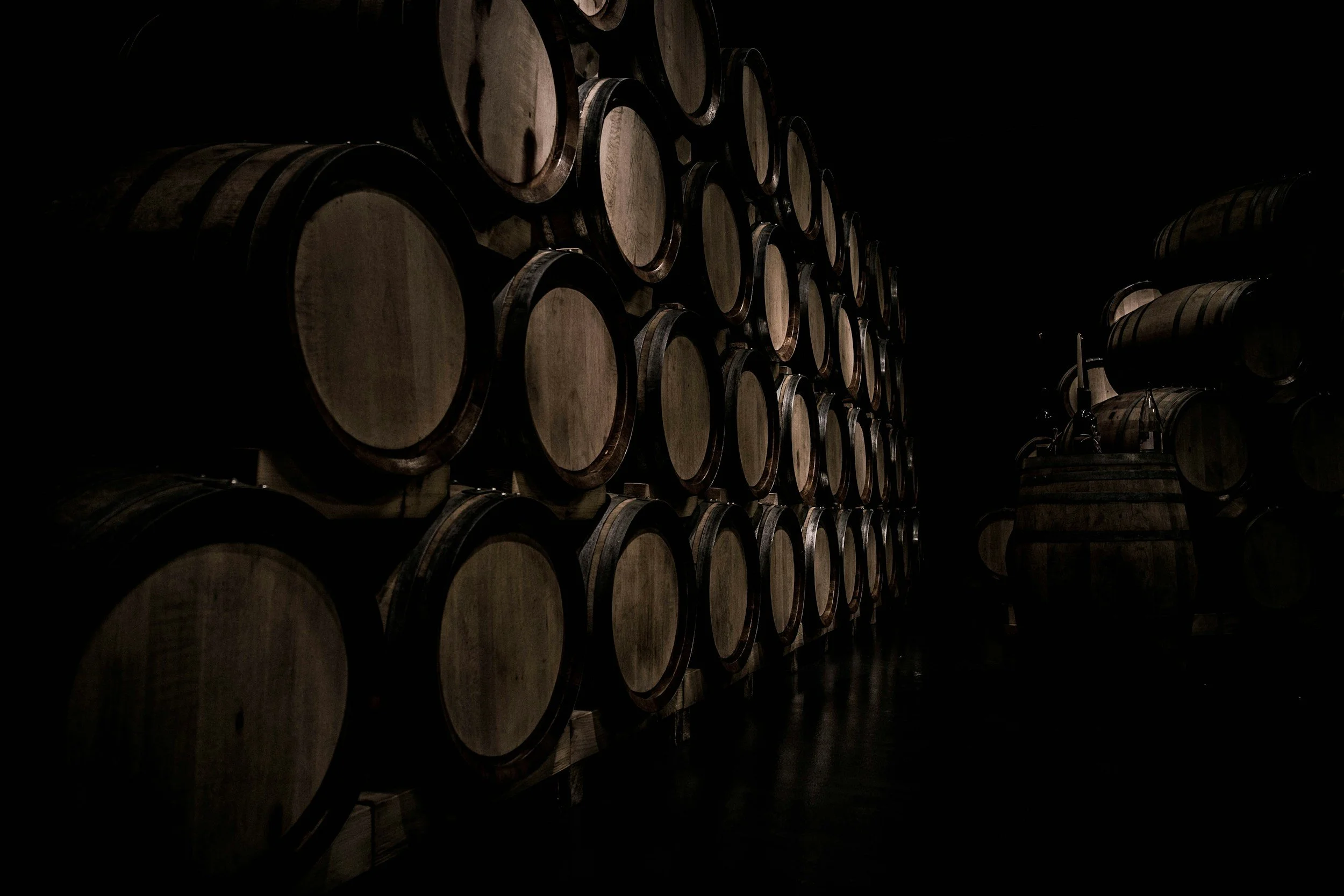 A dark room with stacked wooden barrels, possibly in a wine or bourbon cellar.